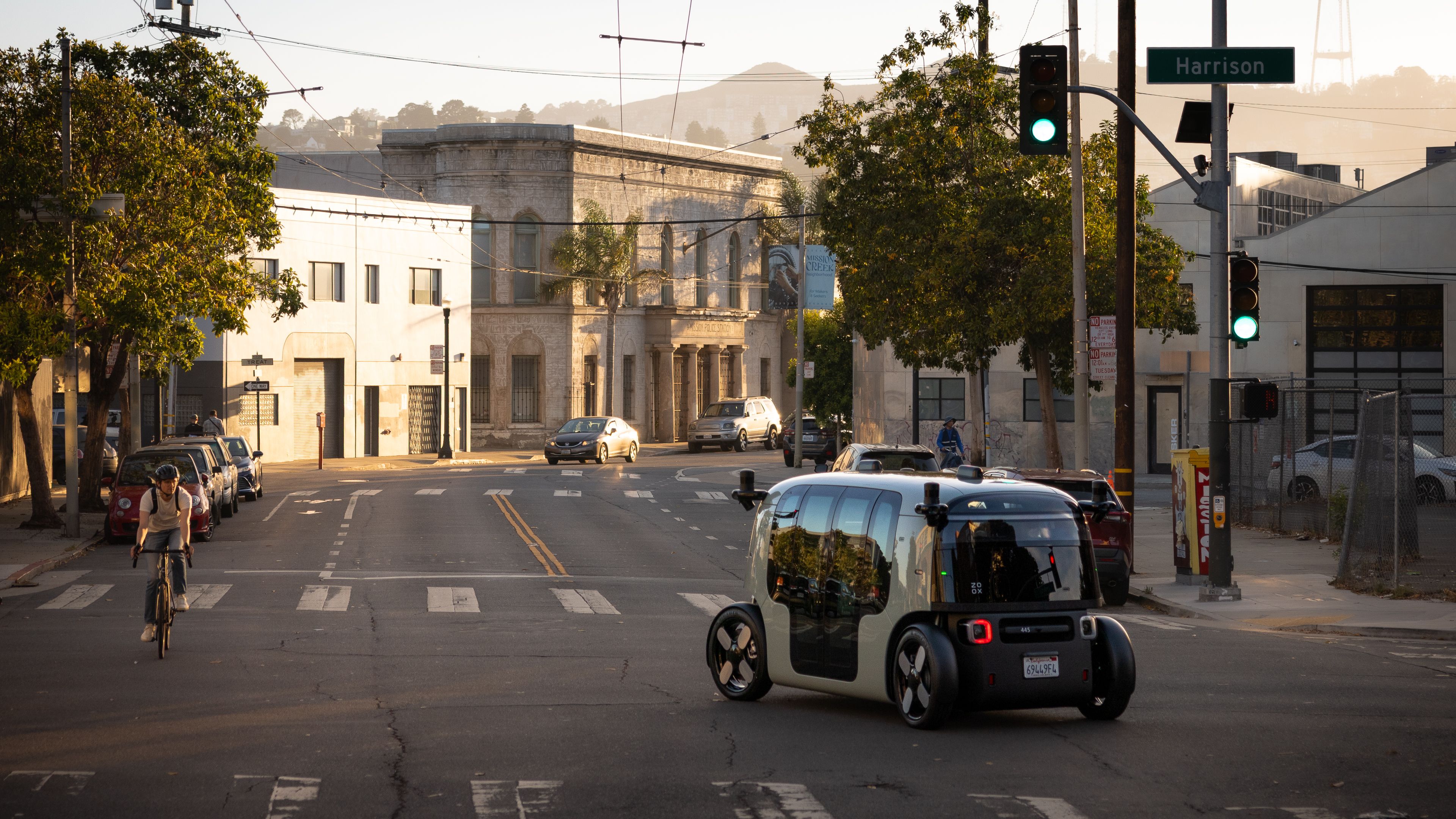 Street view at Harrison with a modern, boxy, black and light gray autonomous vehicle and a cyclist riding toward the camera, under golden sunlight in an urban area.