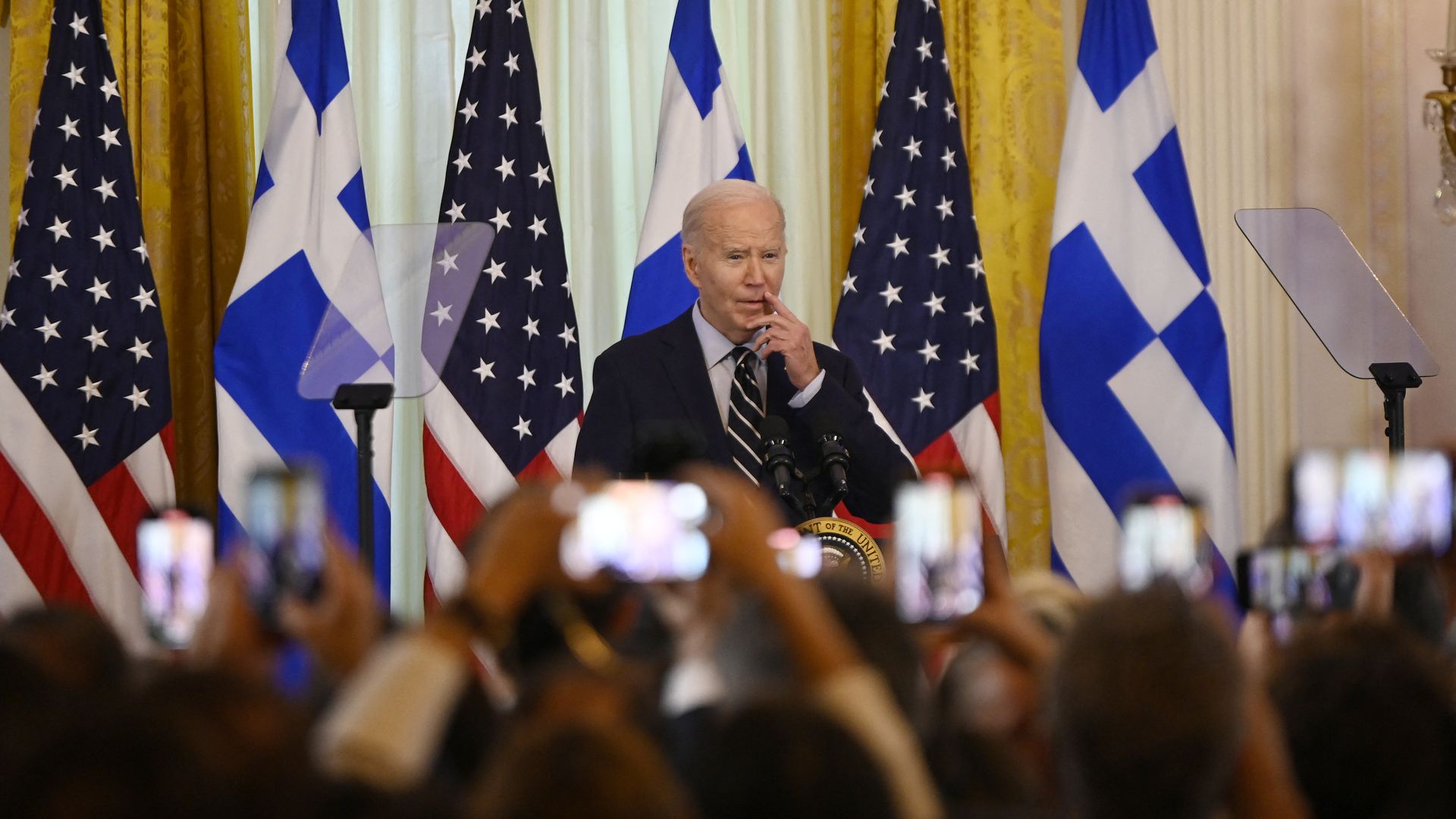 President Biden, wearing a dark gray suit, light blue shirt and blue-and-white striped tie, standing in front of a line of American and Greek flags while the audience takes photos.
