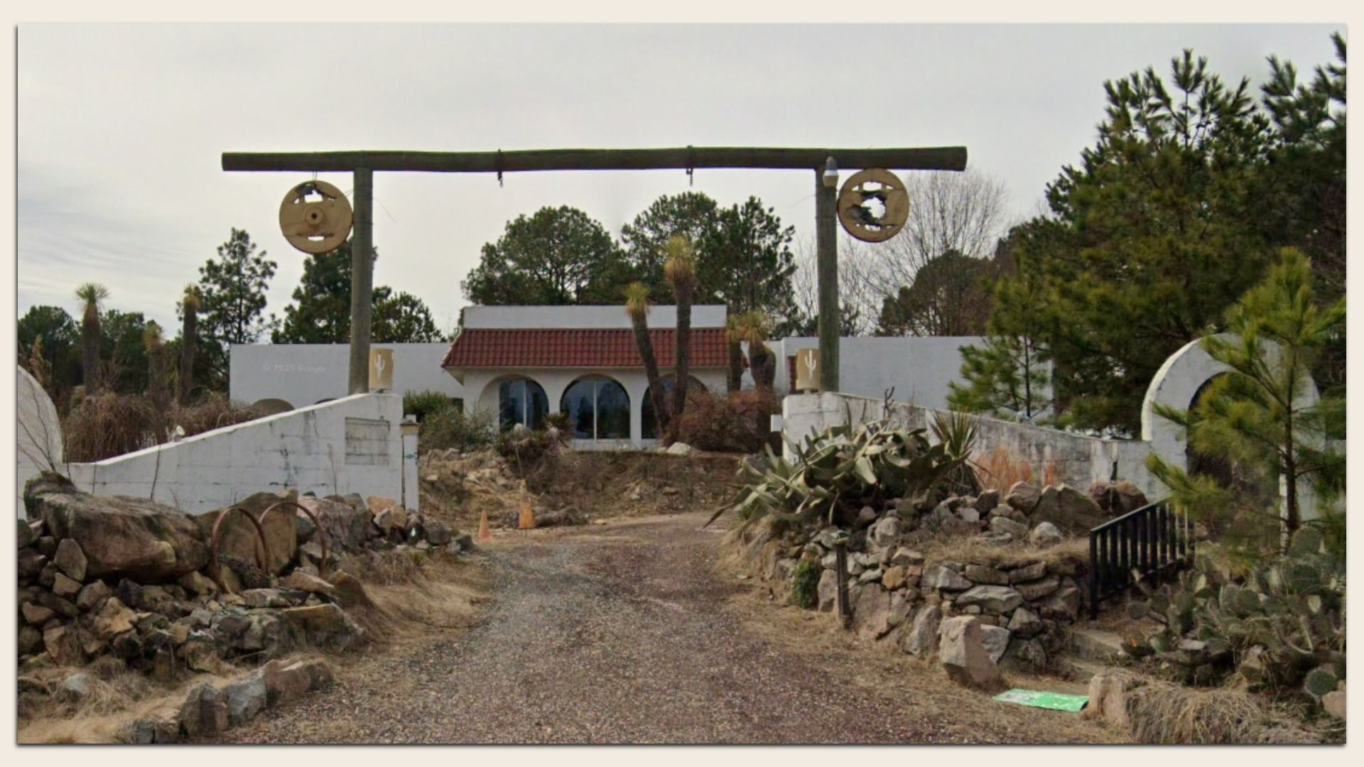 Gravel driveway leads to a white building with a red-tiled roof and arched windows; a large wooden beam gate with circular wagon wheels spans the entrance, flanked by rocks and palm trees.