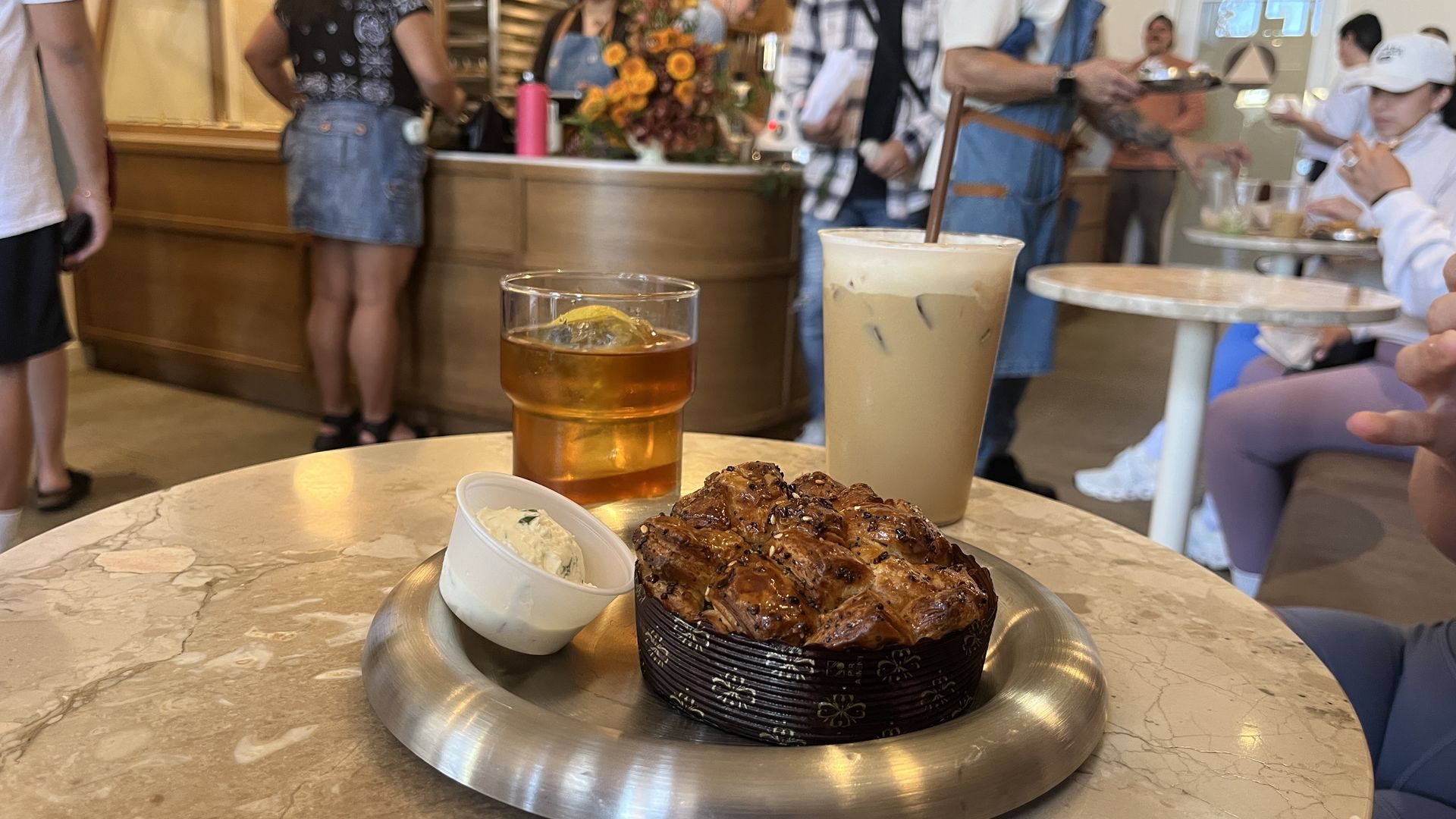A glazed pastry, a small cup of cream cheese, a glass of a coffee drink that looks like iced tea with lemon, and a tall iced latte sit on a marble table. In the background, cafe staff and customers stand at the counter.