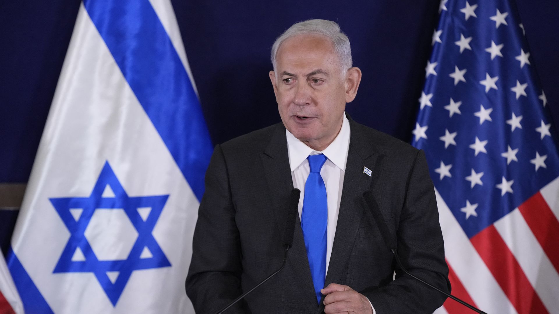 Israeli Prime Minister Benjamin Netanyahu, wearing a dark gray suit, white shirt and blue tie, standing between U.S. and Israeli flags.