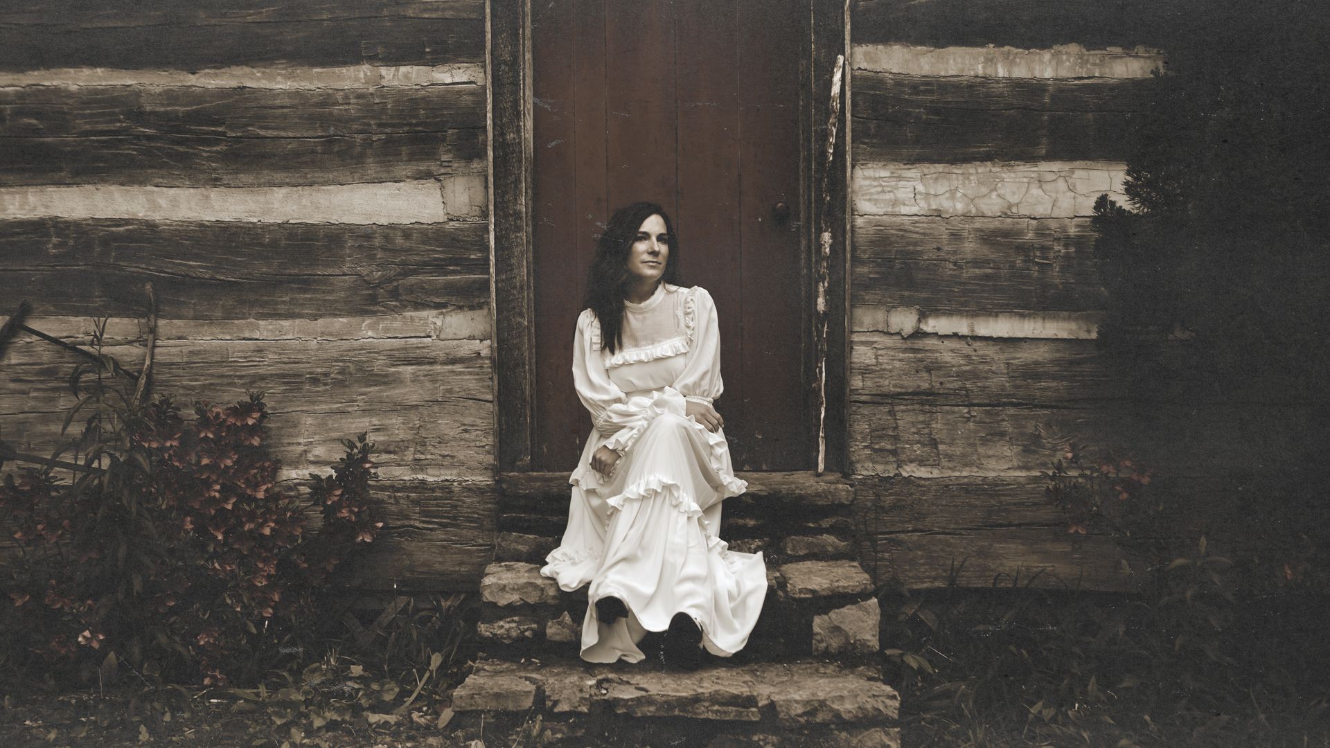 A woman in a long white vintage dress sits on stone steps before a weathered log cabin door, with overgrown plants and a muted sepia tone.