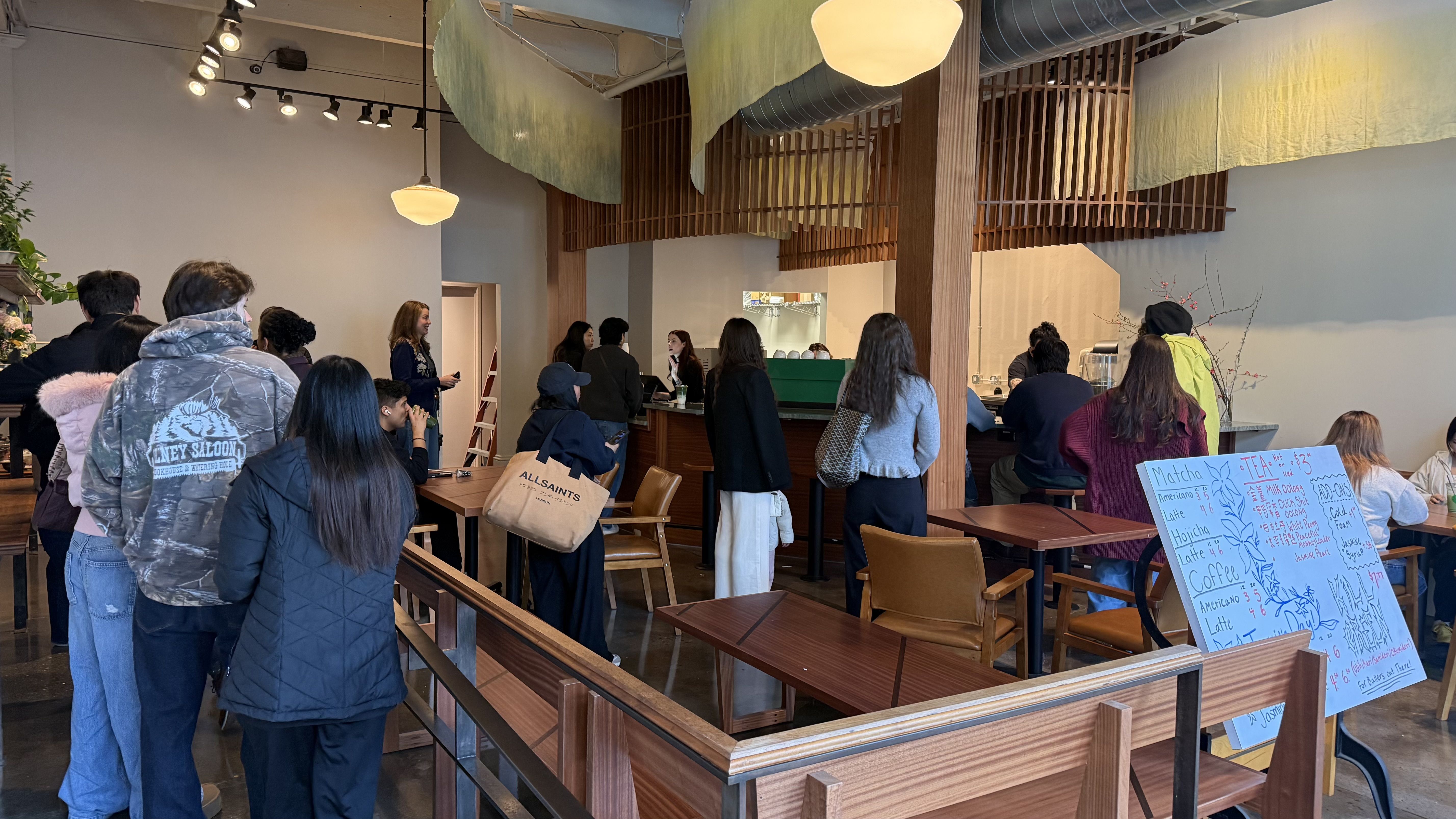People standing in line and sitting at tables inside a modern coffee shop with wooden decor and hanging green fabric. A handwritten menu board lists drinks and prices.