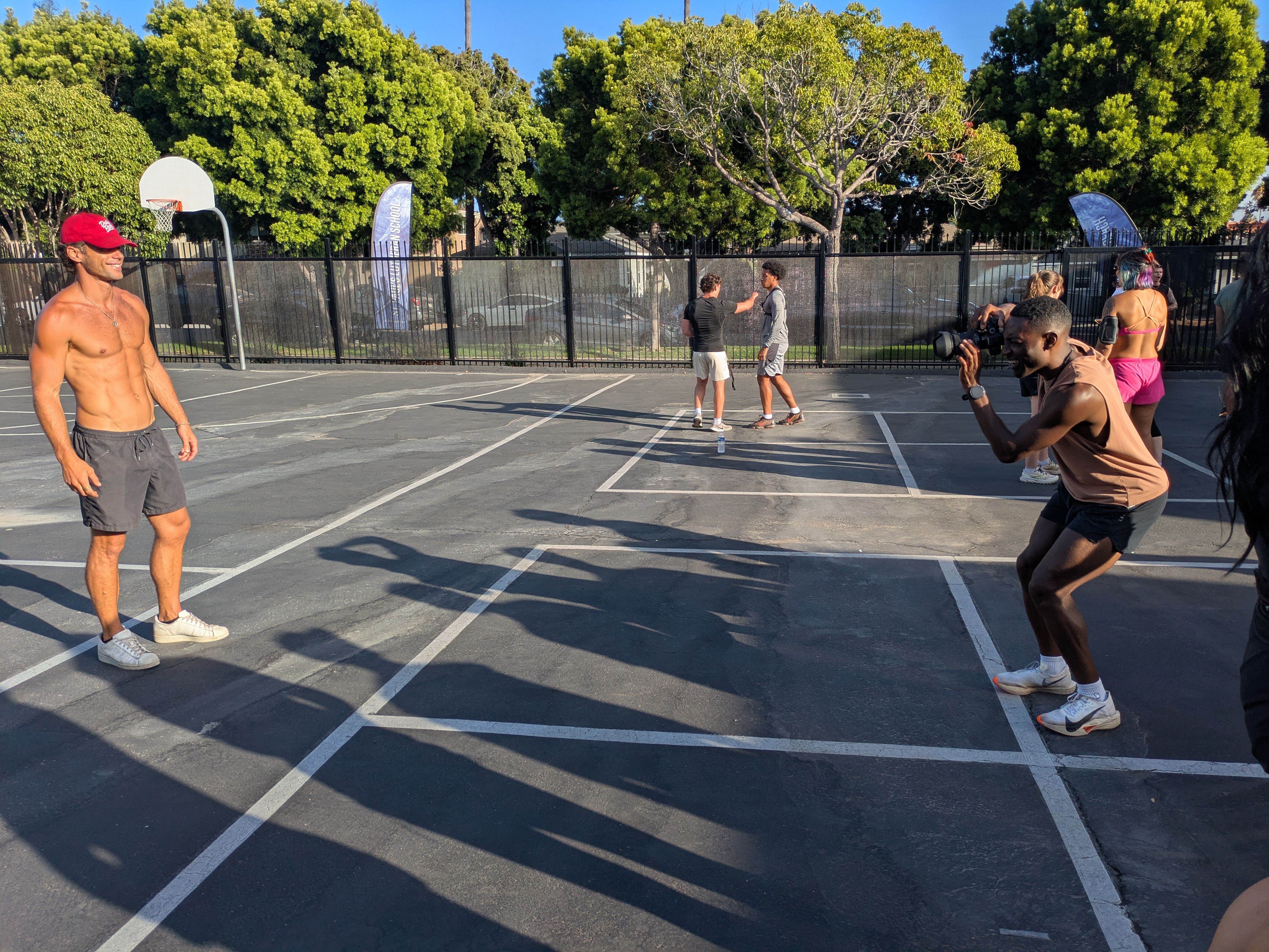 Outdoor basketball court with a shirtless man in a red cap on the left, two men chatting near the fence center, and a cameraman filming a group on the right, with green trees and blue sky behind.
