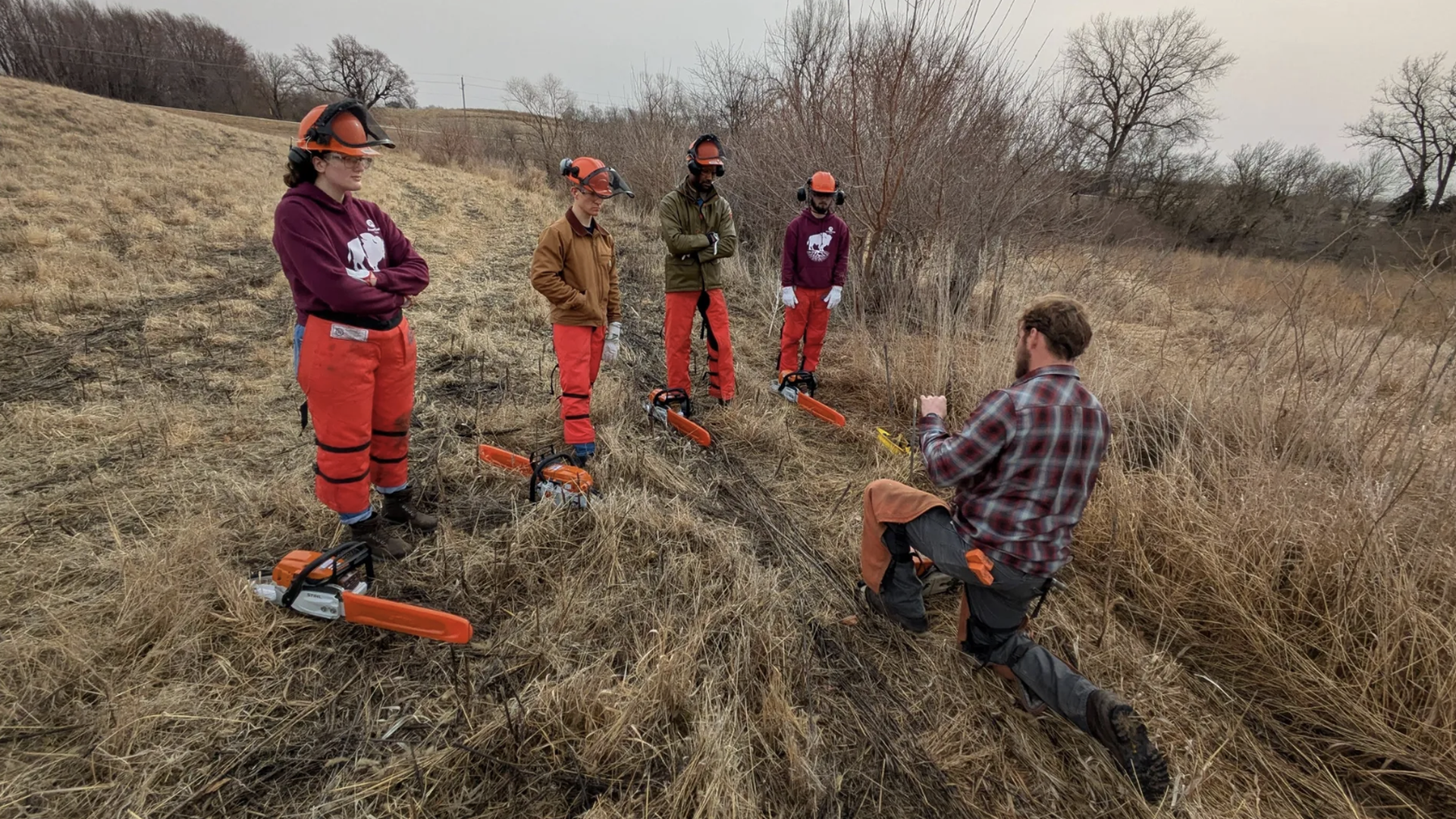 Five students in orange pants and orange helmets stand in a dry grassy field, listening to an instructor in a plaid shirt kneeling and showing a yellow tool; orange chainsaws rest on the grass.