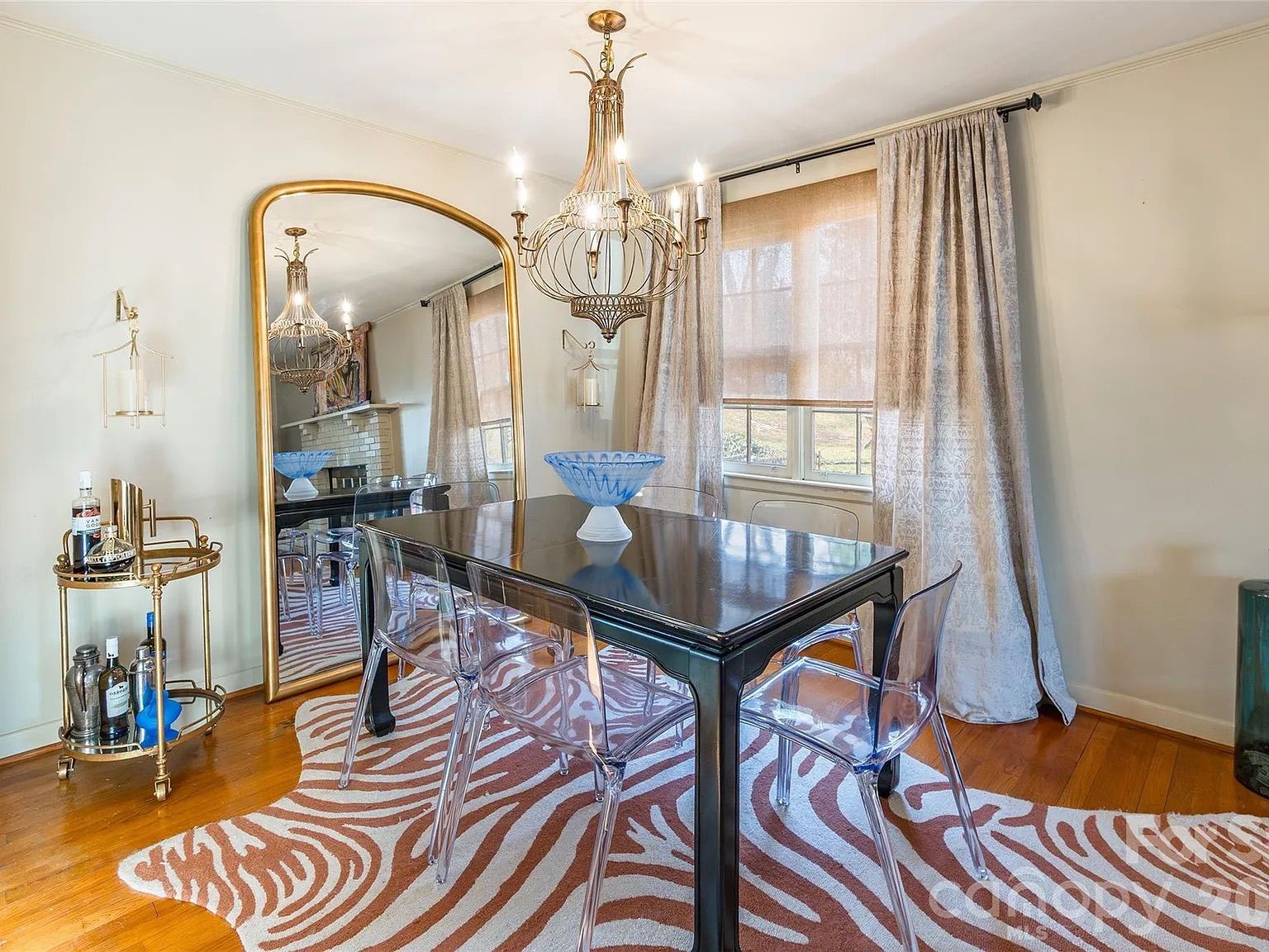 Dining room with a black table, four clear chairs, a large gold-framed mirror, blue centerpiece bowl, gold chandelier, patterned beige curtains, and a zebra-patterned rug on wooden floor.