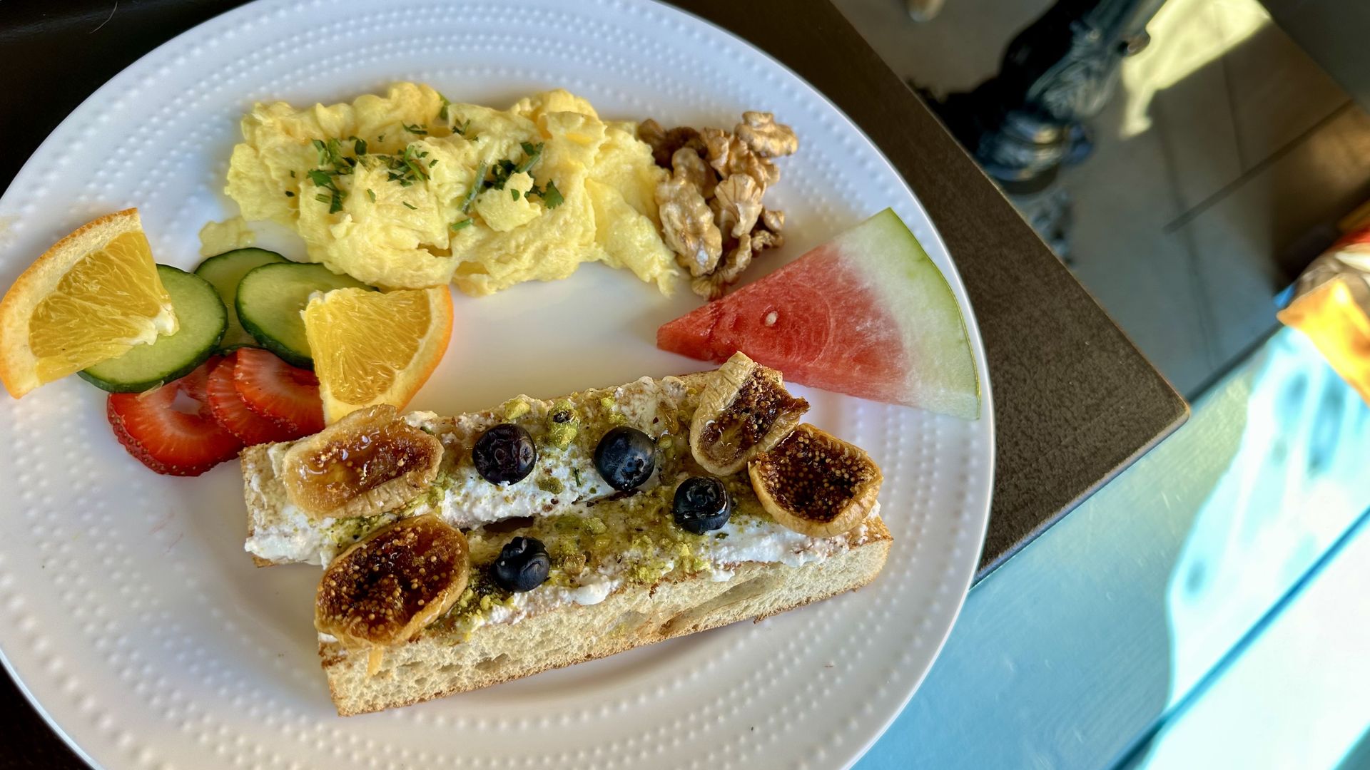 A photo of a plate with fig and cheese toast, scrambled eggs and sliced pieces of fruit. 
