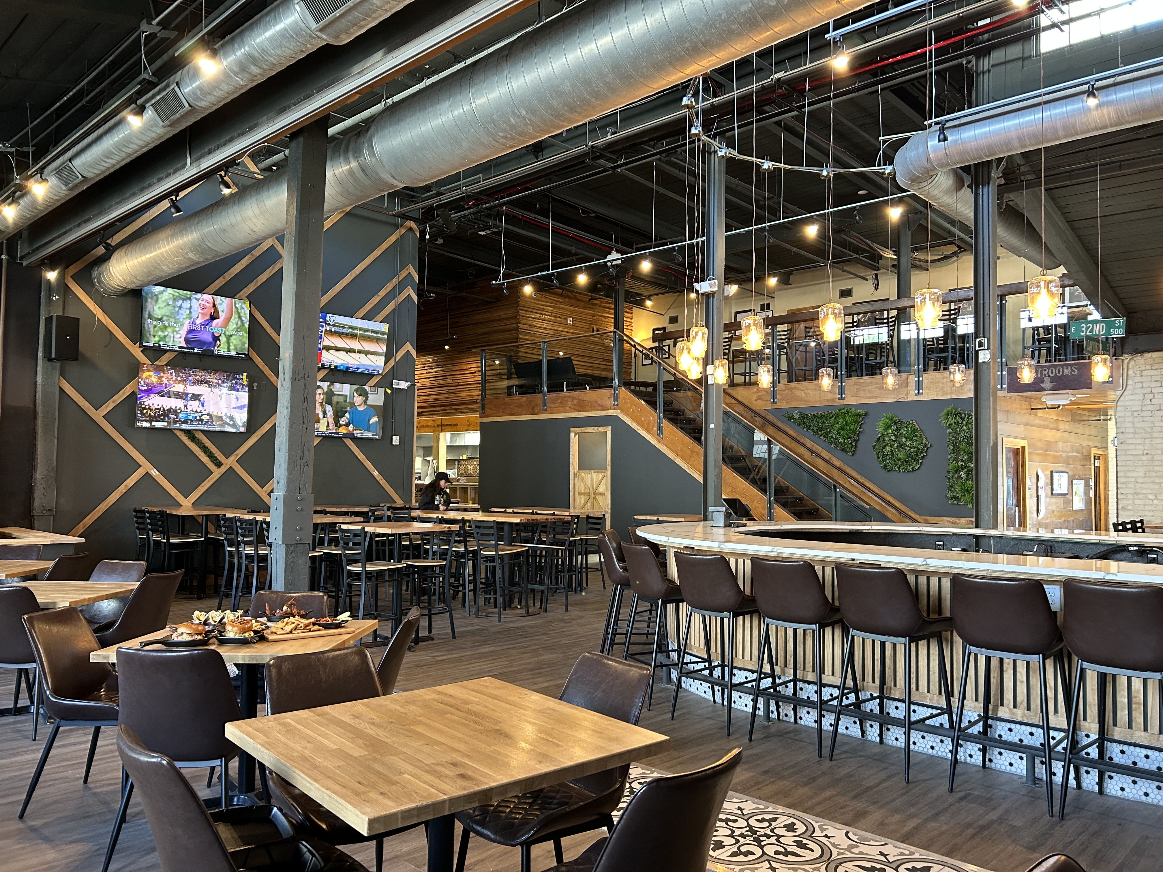 Heist Brewery NoDa. Industrial cafe interior with exposed silver ducts, hanging bulbs, and wood-paneled walls. Tables and brown chairs fill the floor; a mezzanine with stools overlooks the space, pastries on a table.