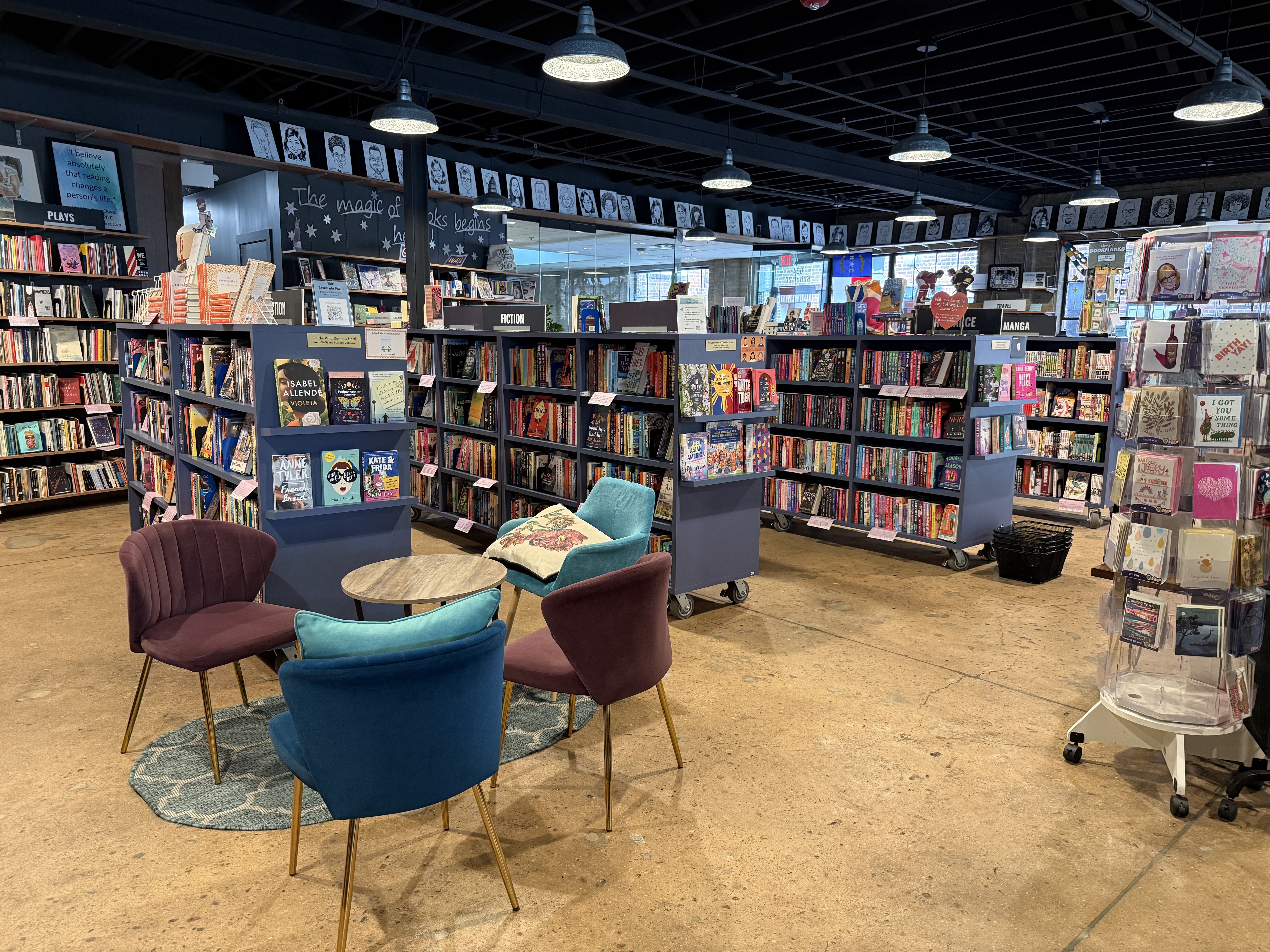 Interior of a bookstore: tall shelves of colorful books, round tables with blue and pink chairs, hanging pendant lights, and a chalkboard sign that reads "The magic of books begins here".