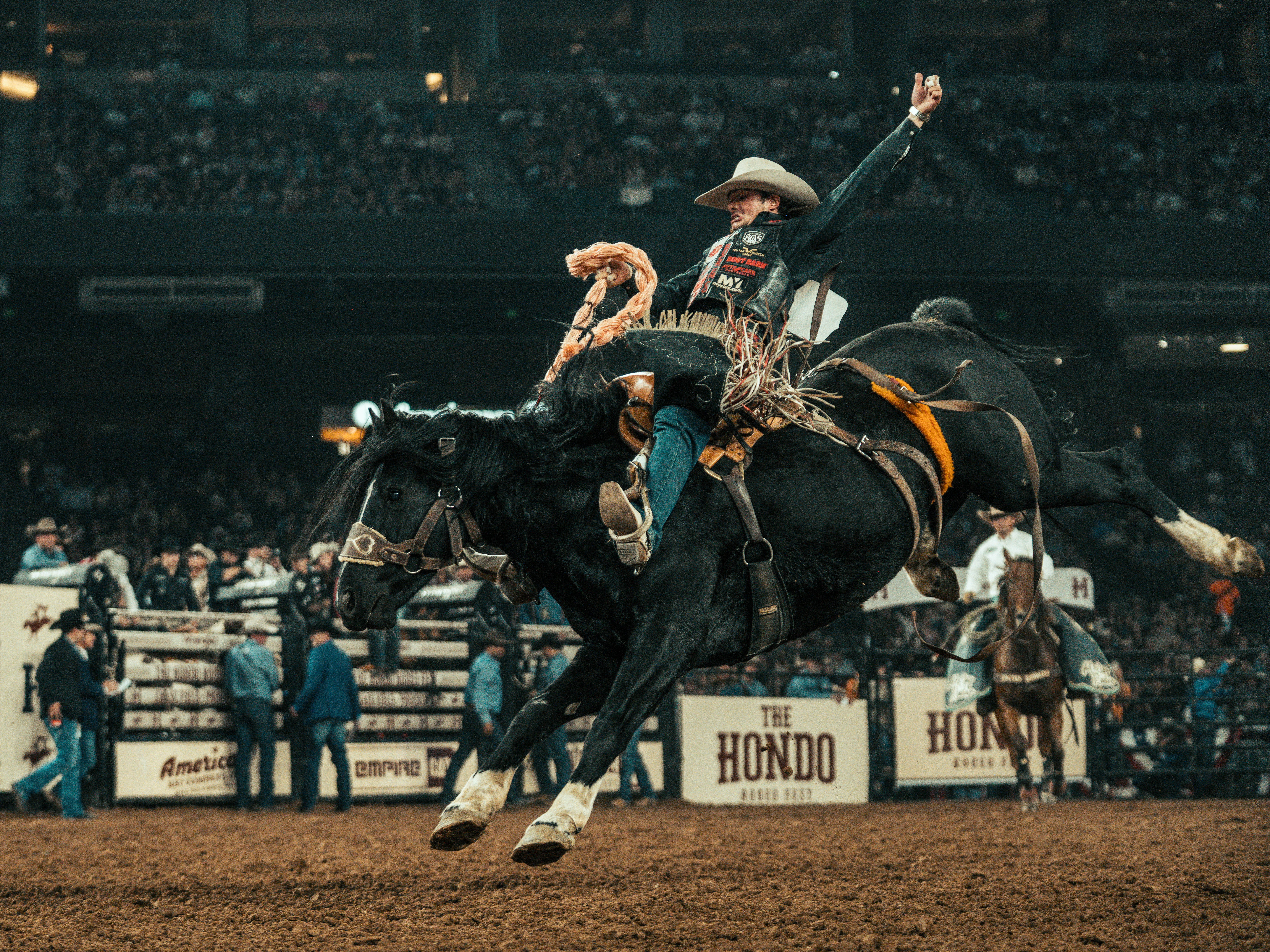 A cowboy rides a bucking black horse mid-air in a crowded rodeo arena. He wears a beige hat, dark vest, jeans, and boots, with a rope in one hand; sponsor banners line the rails in the background.