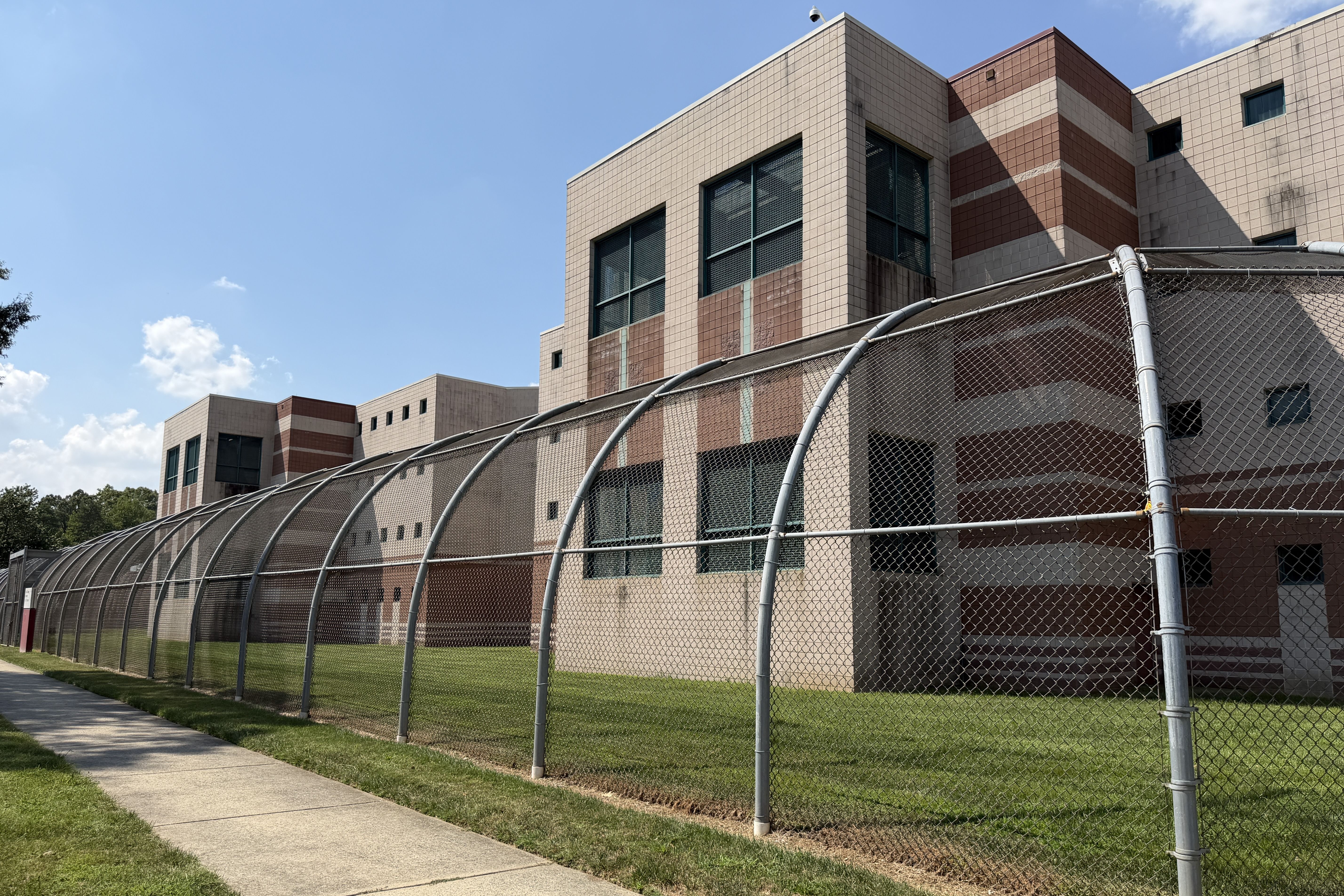 Beige and brown tiled building with large grid windows rises behind a curved chain-link fence; a security camera sits atop the roof while a blue sky and green grass line the sidewalk on the left.