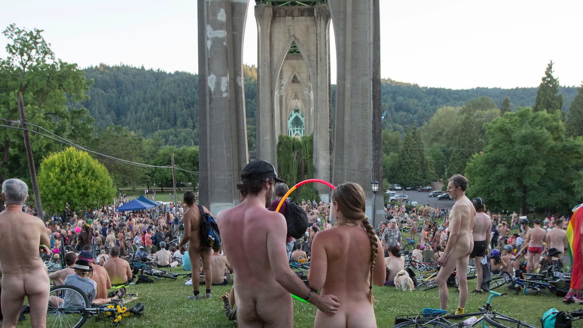 Large group of naked people gathered on a grassy area under a concrete bridge, surrounded by green trees and hills, with many bicycles scattered around.