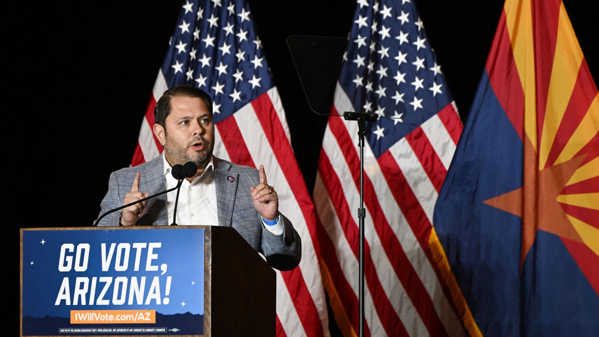 A man standing at a  podium in front of American and Arizona flags.