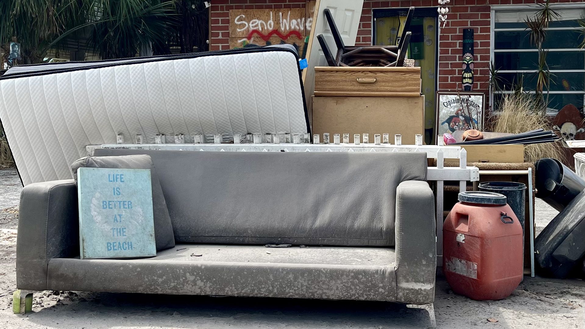 A gray couch frame, mattress and other home items sit in front of a red brick house. On the couch, a sign reads, "Live is better at the beach." 