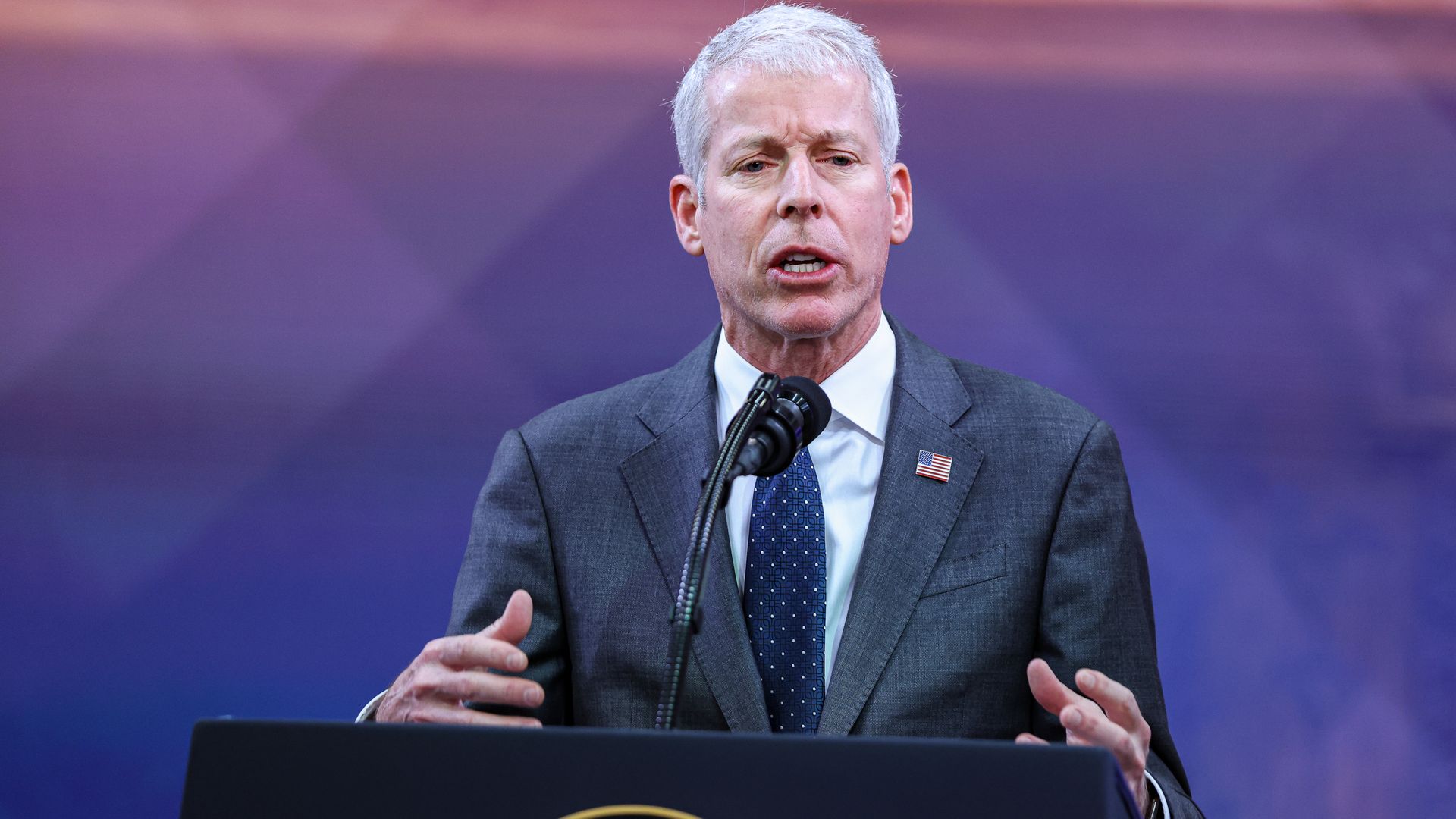 A person speaks at a microphone in a suit and tie