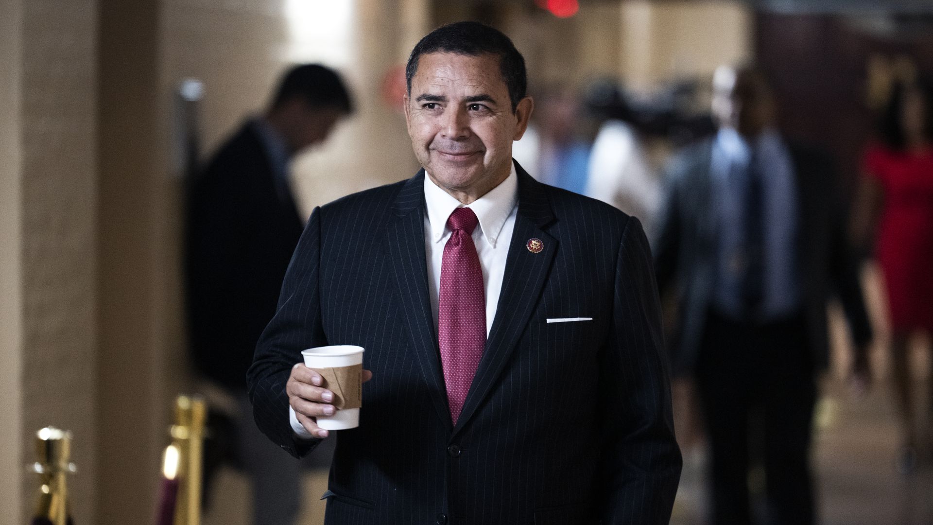 Rep. Henry Cuellar, wearing a pinstripe suit, white shirt and red tie and holding a cup, walking through a Capitol basement hallway.