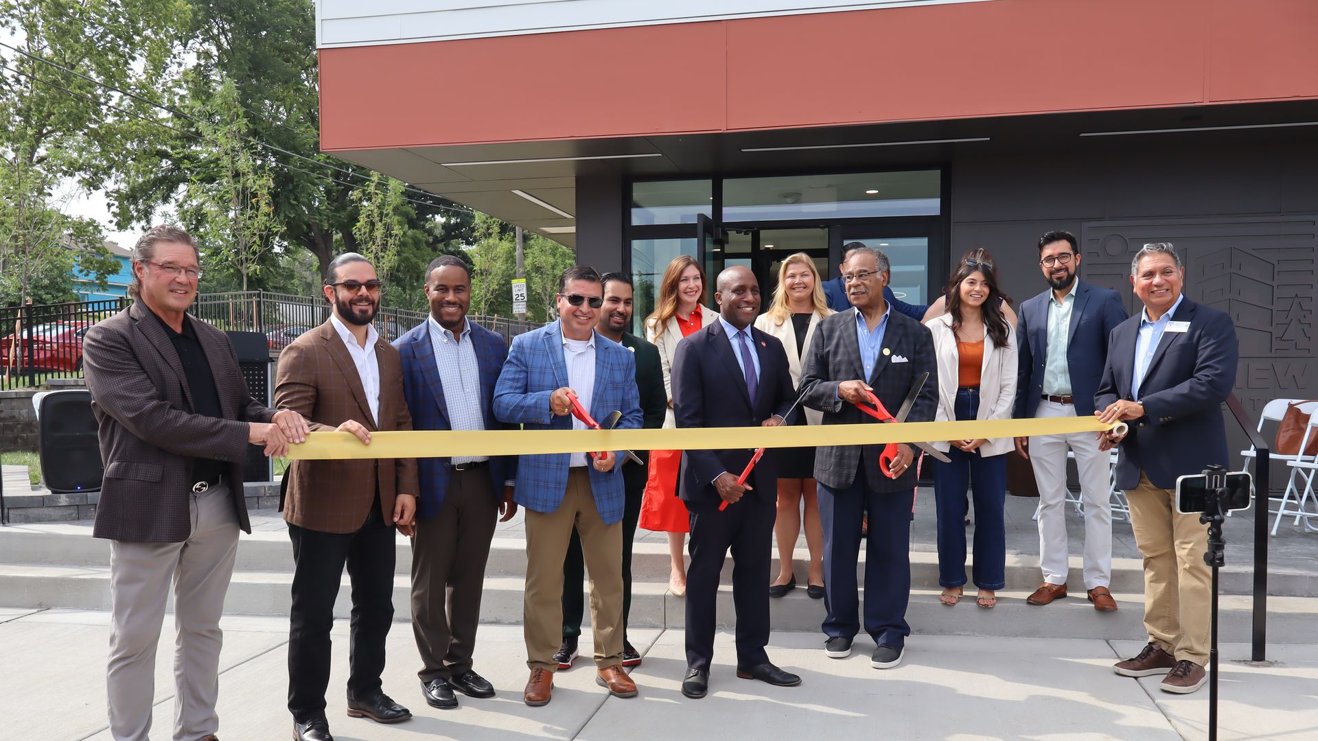 Group of diverse people in business casual attire standing outside building during ribbon-cutting ceremony, holding large yellow ribbon and oversized red scissors.