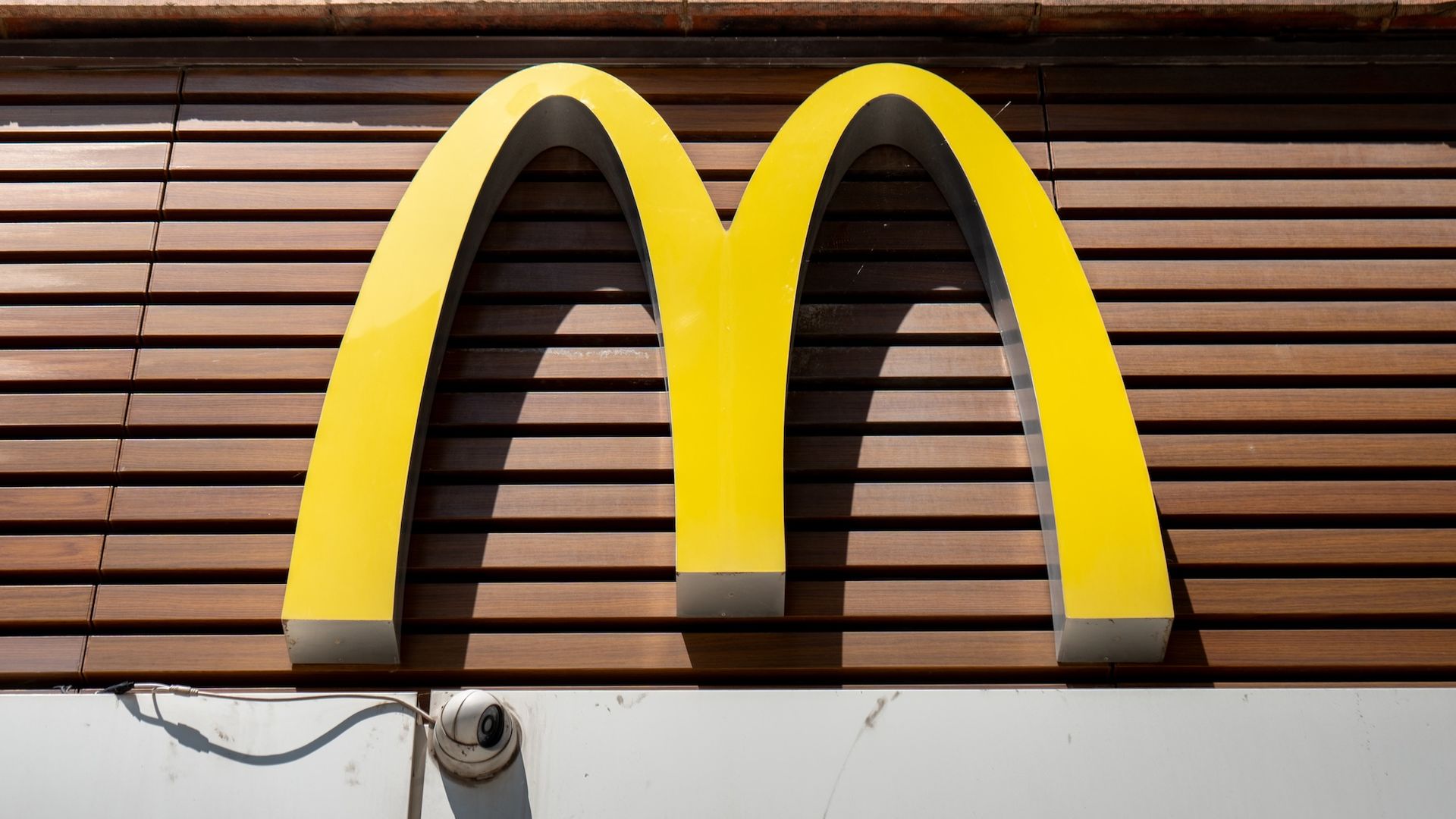 Close-up of the bright yellow McDonald's golden arches logo mounted on a brown wooden slat facade, with a white lower wall and exposed pipes below.