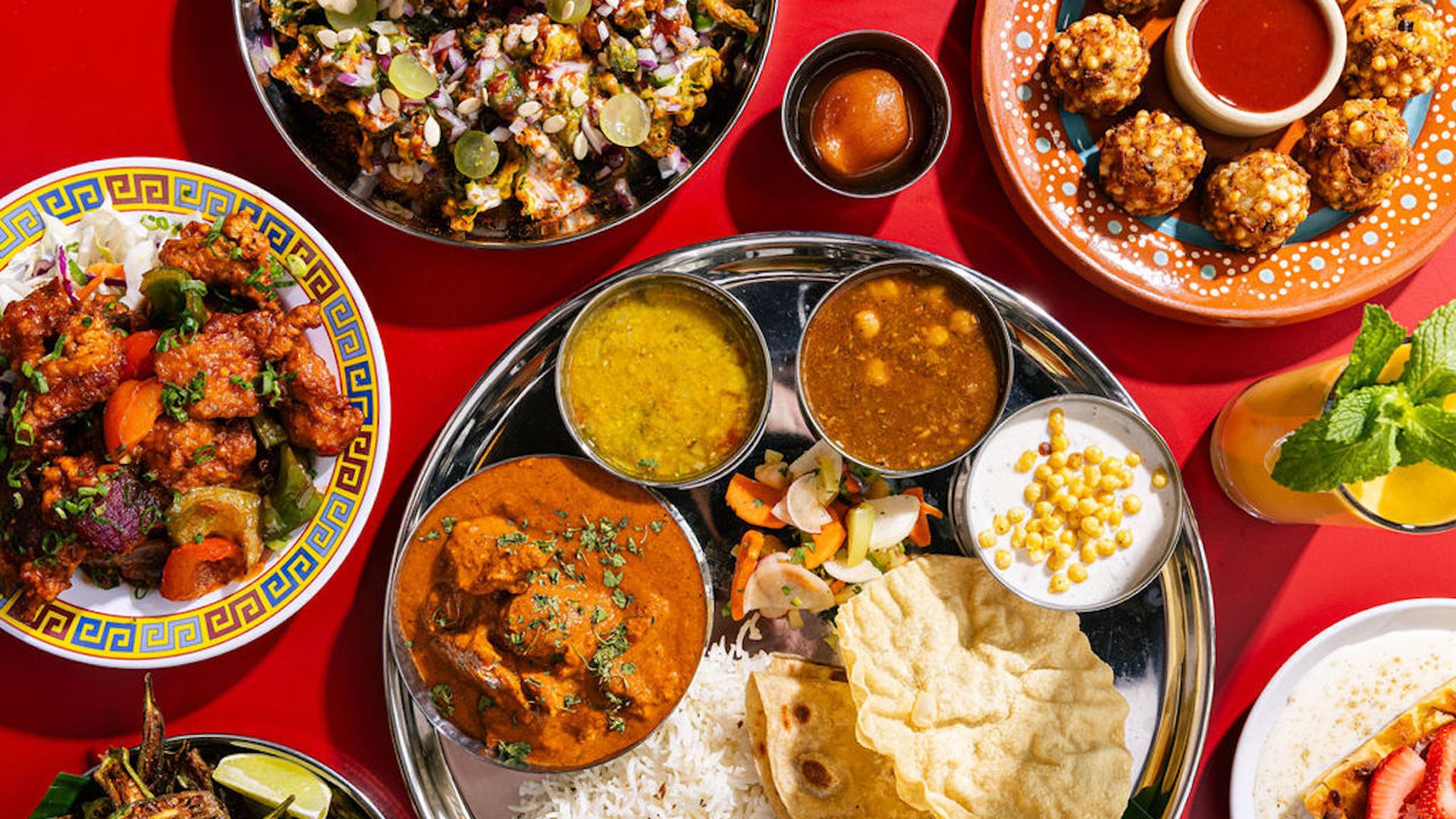 Indian meal on a red table with a thali plate featuring rice, curry, dal, chapati, papad, and chutneys, surrounded by dishes of fried okra, vegetable stir-fry, snack balls, and a mango drink garnished with mint.
