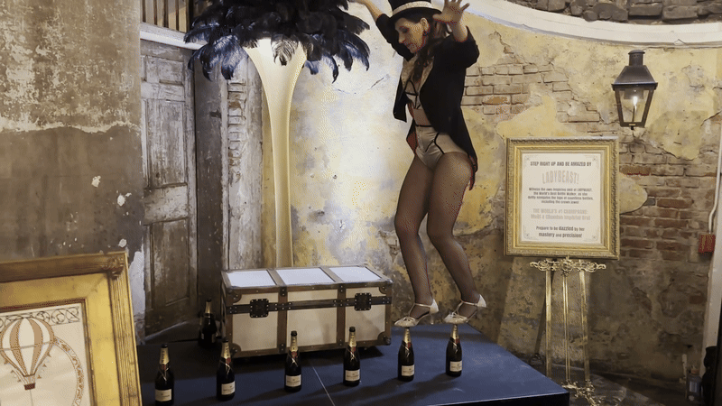 A performer dressed in black and white vintage attire balances on champagne bottles on a stage with rustic brick walls, a feather decoration, and a framed sign nearby.