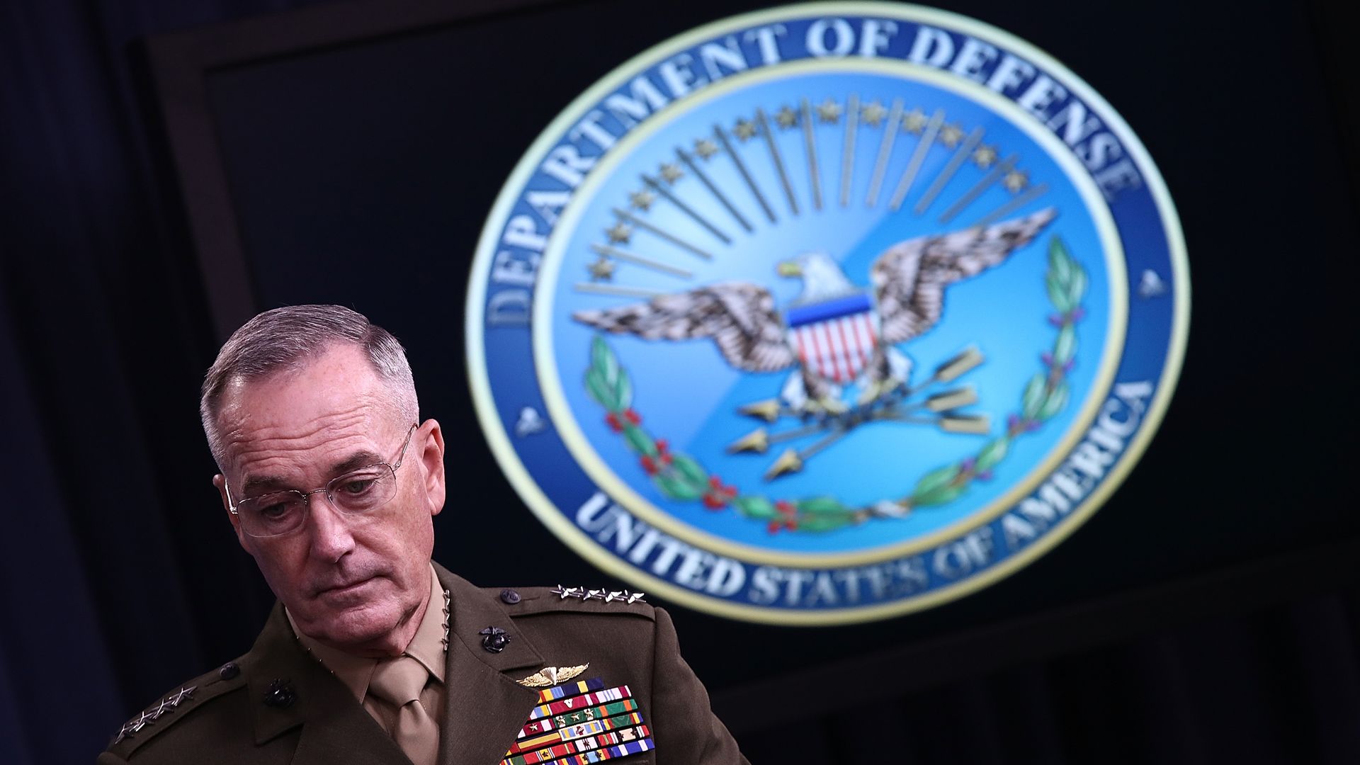 Joseph Dunford, in military uniform, stands in front of a large Defense Department seal. The background is dark.