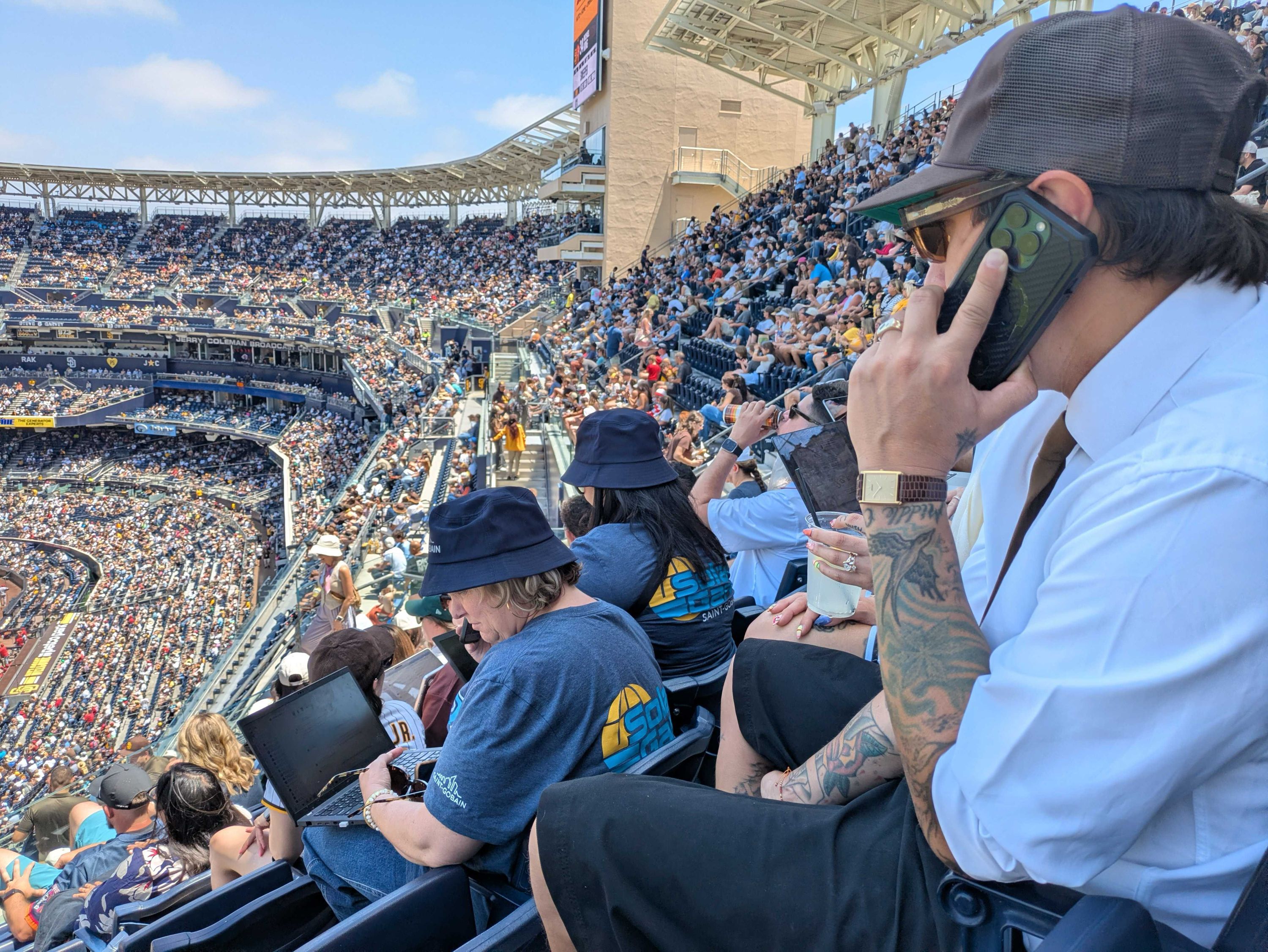 Crowded baseball stadium under a clear sky. In the foreground a man in a light shirt and brown cap talks on a large smartphone, showing tattoos; others use laptops.