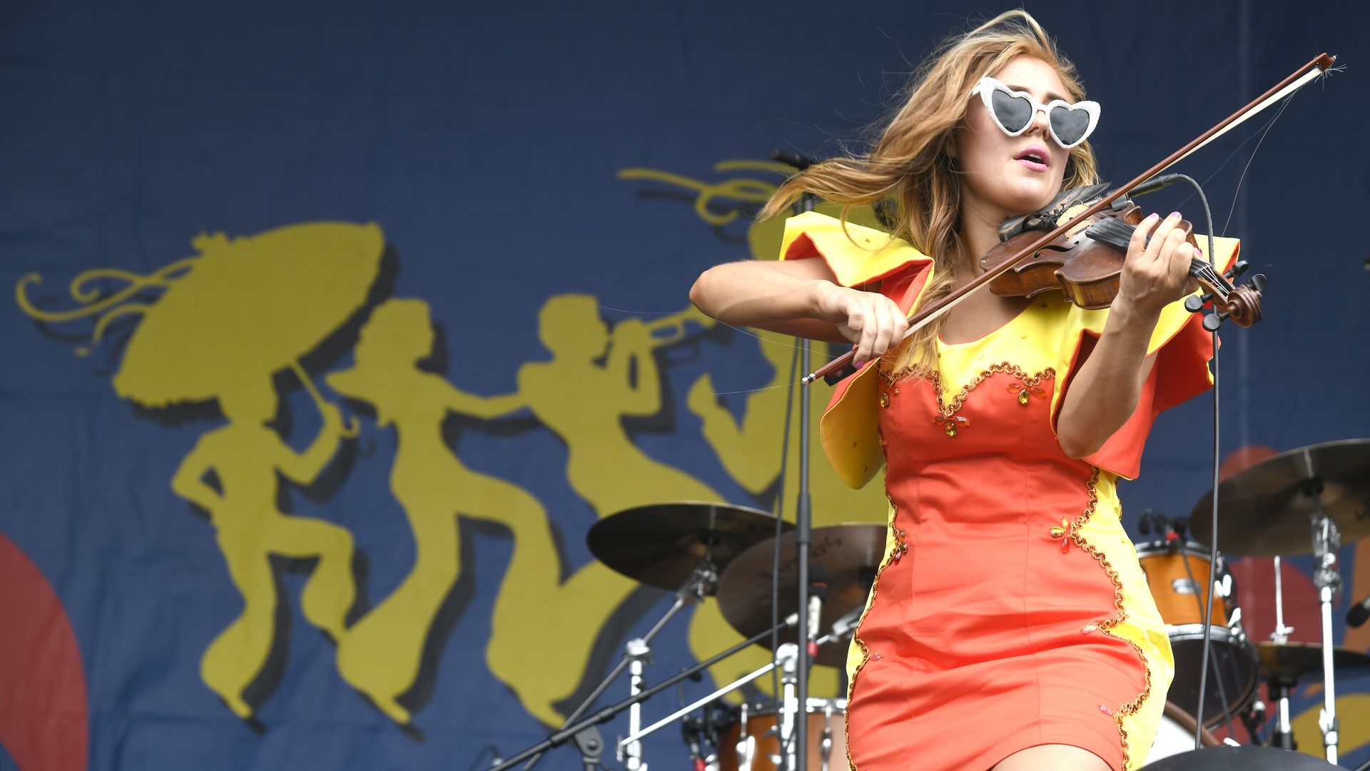 A woman wearing heart-shaped sunglasses plays a fiddle in a yellow and orange dress. The Jazz Fest logo is on a stage background behind her.