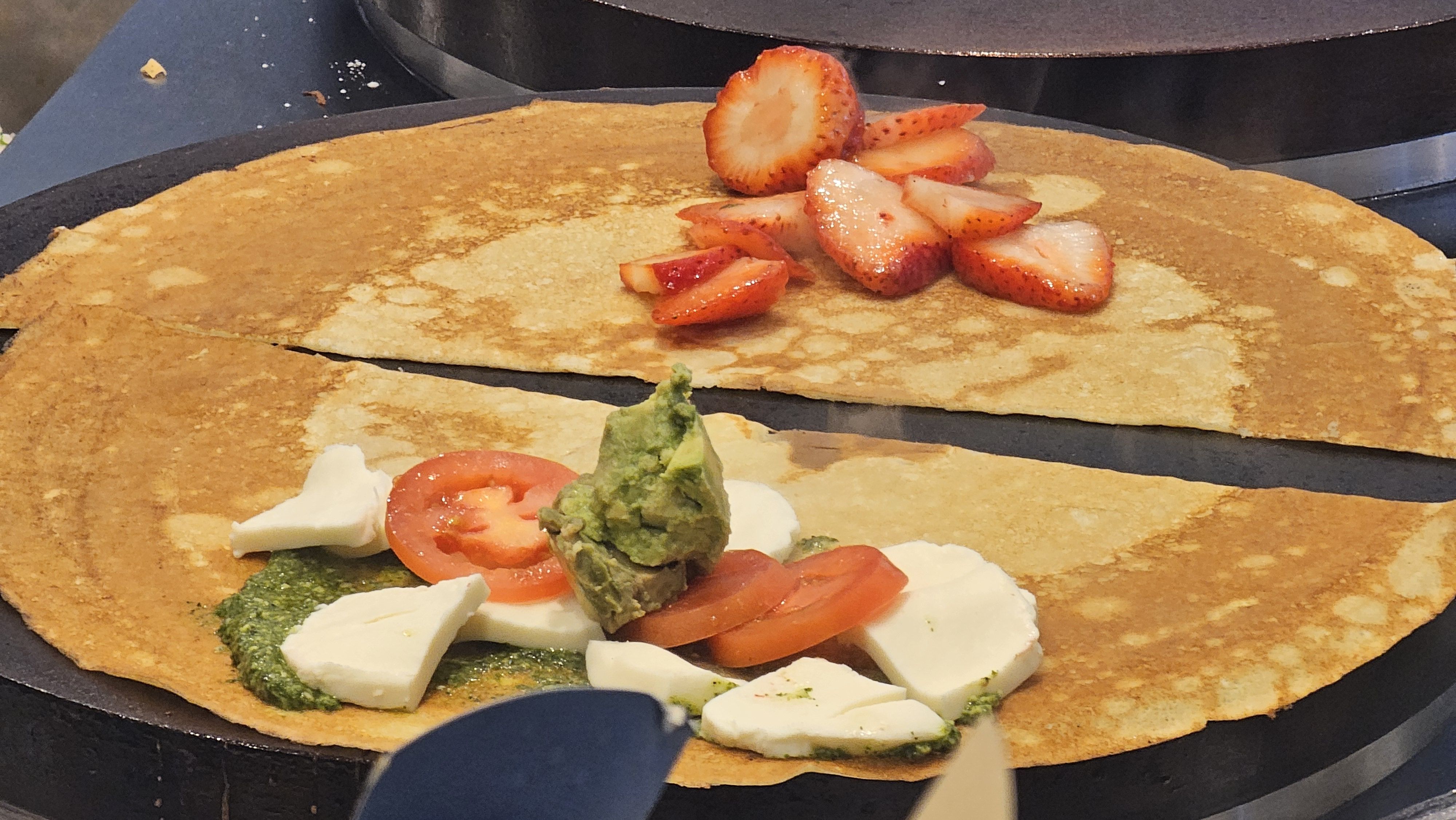 Two crepes on a griddle; one topped with sliced strawberries, the other with tomato slices, mozarella cheese, pesto, and avacado.