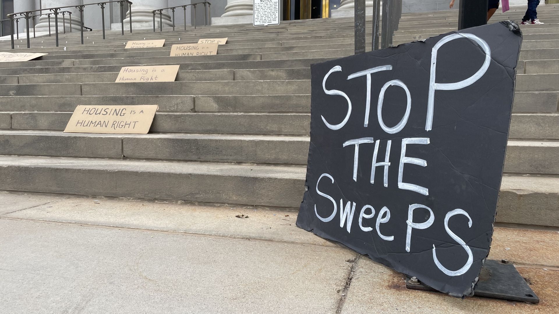 A handwritten sign that reads STOP THE SWEEPS near several steps with more signs saying HOUSING IS A HUMAN RIGHT. 