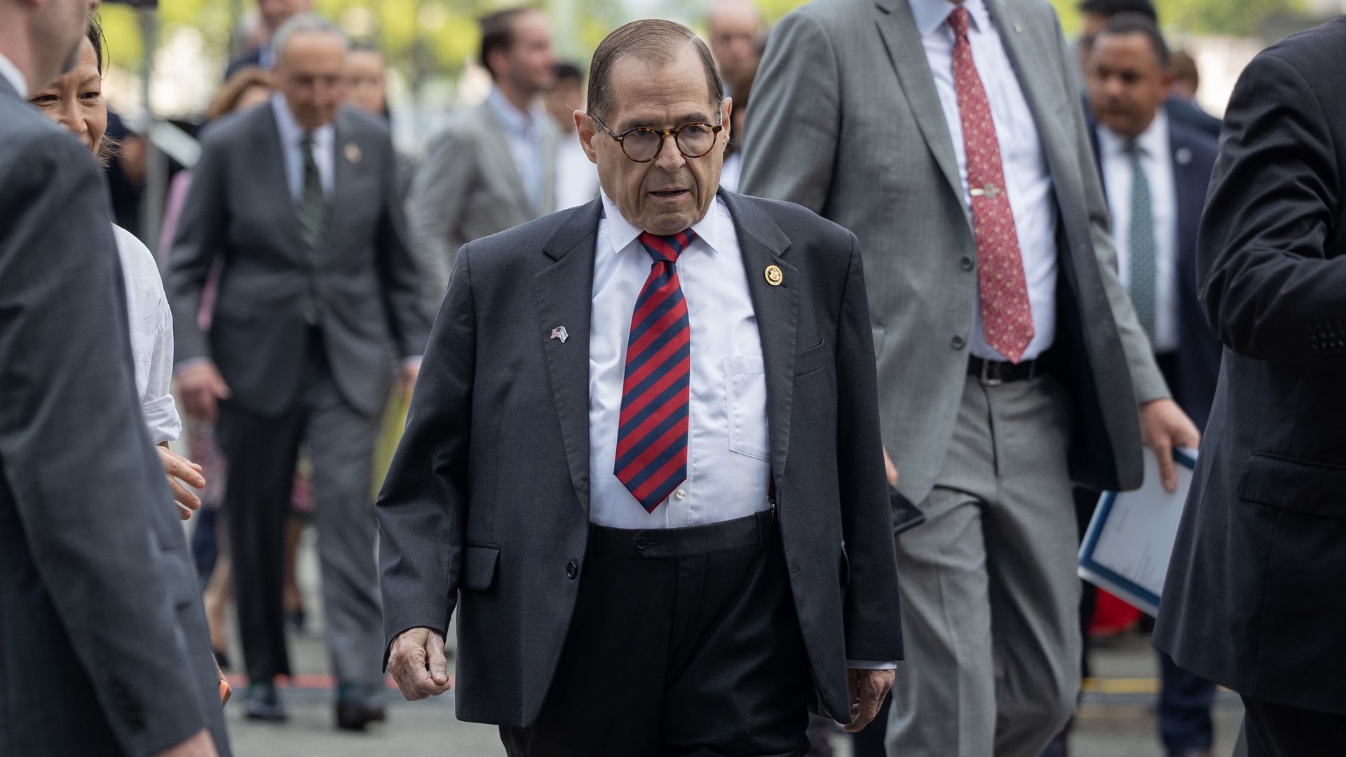 Man in dark suit jacket, white shirt, and red-striped tie walking among a crowd of people, some in suits, on a city street during daytime.