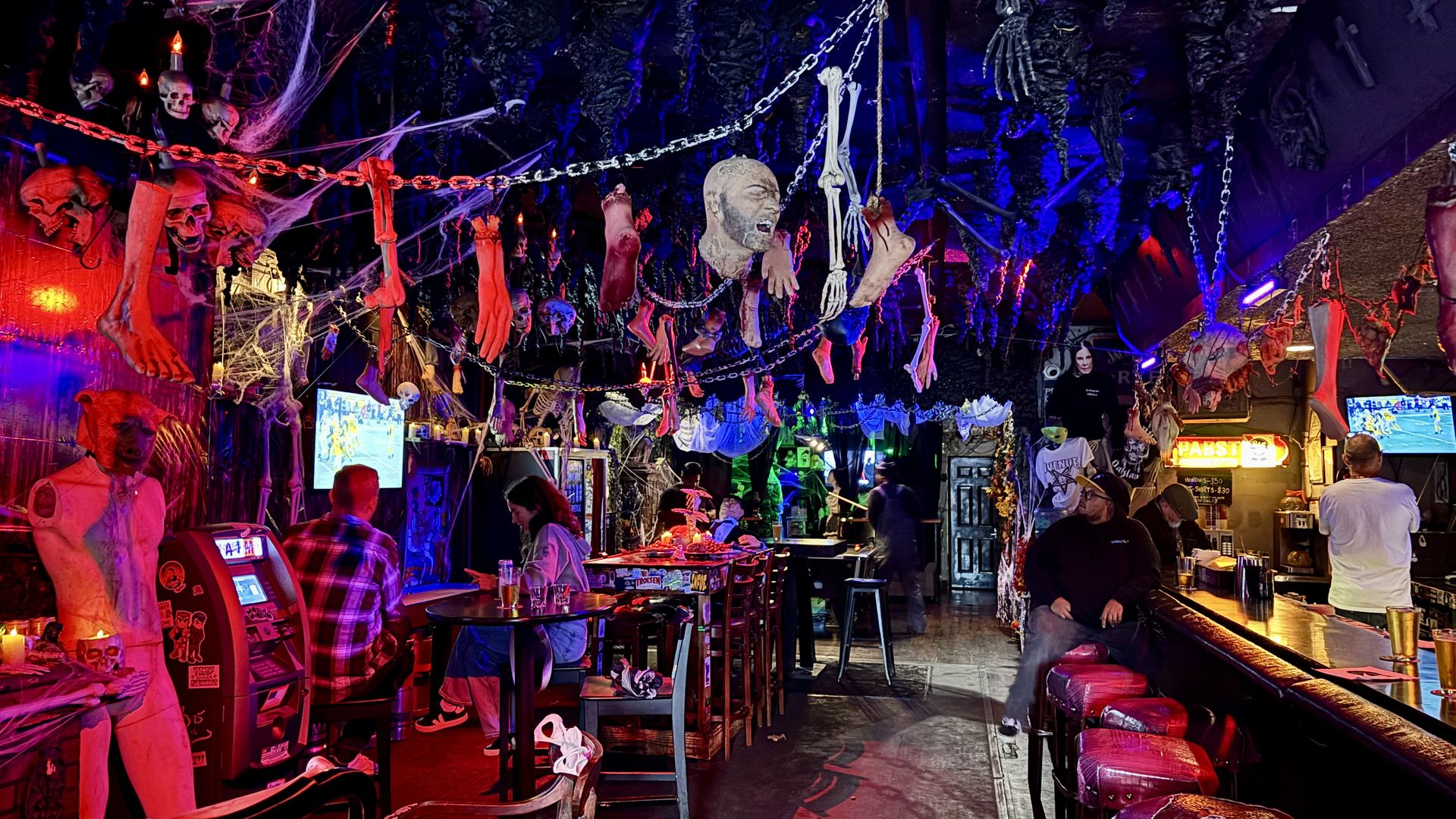 Interior of a dimly lit bar decorated with hanging fake limbs, skulls, chains, and cobwebs, creating a spooky atmosphere with red and blue lighting and patrons sitting and watching TV screens.