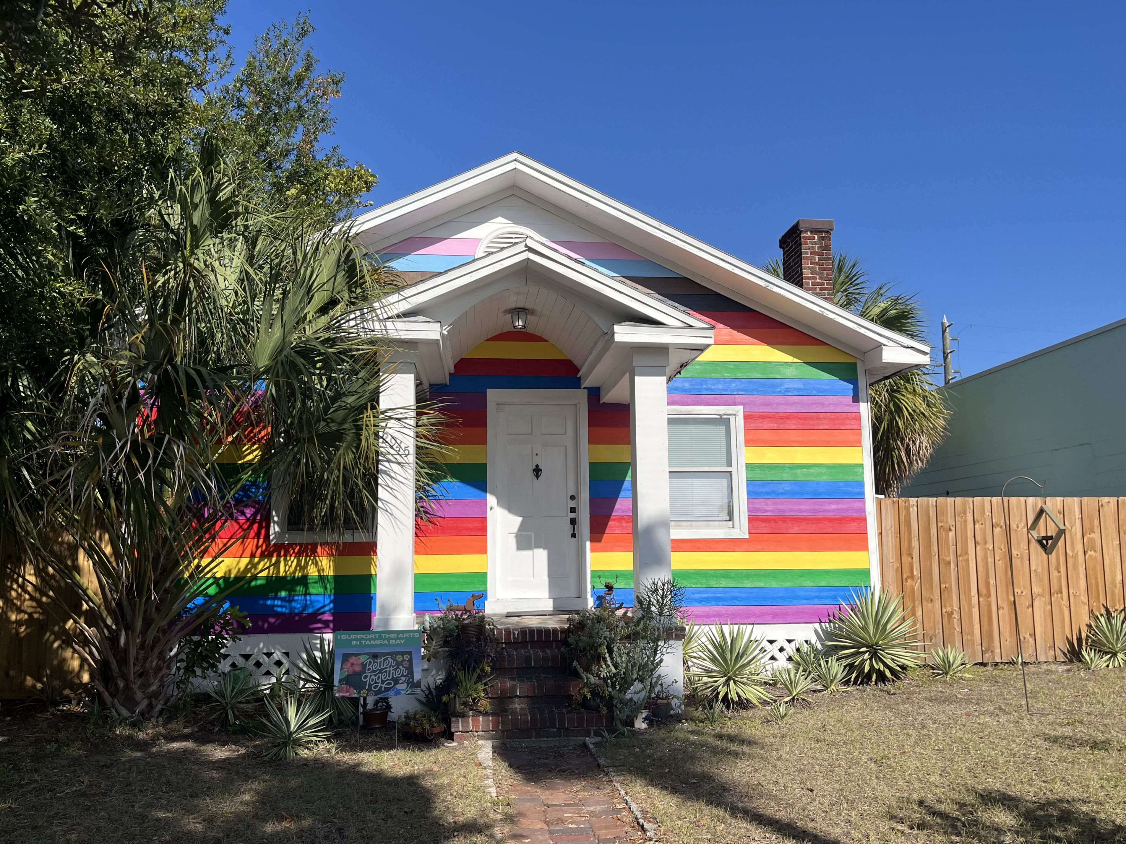 Small house painted with horizontal rainbow stripes, white columns, door, and window. Palm trees and succulents in front yard under clear blue sky.