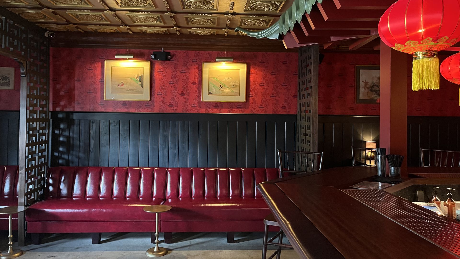 Interior of a Chinese-style bar with ornate gold ceiling tiles, dark wood paneling, red velvet booths along a wall, framed art, and a large red lantern hanging near the bar.
