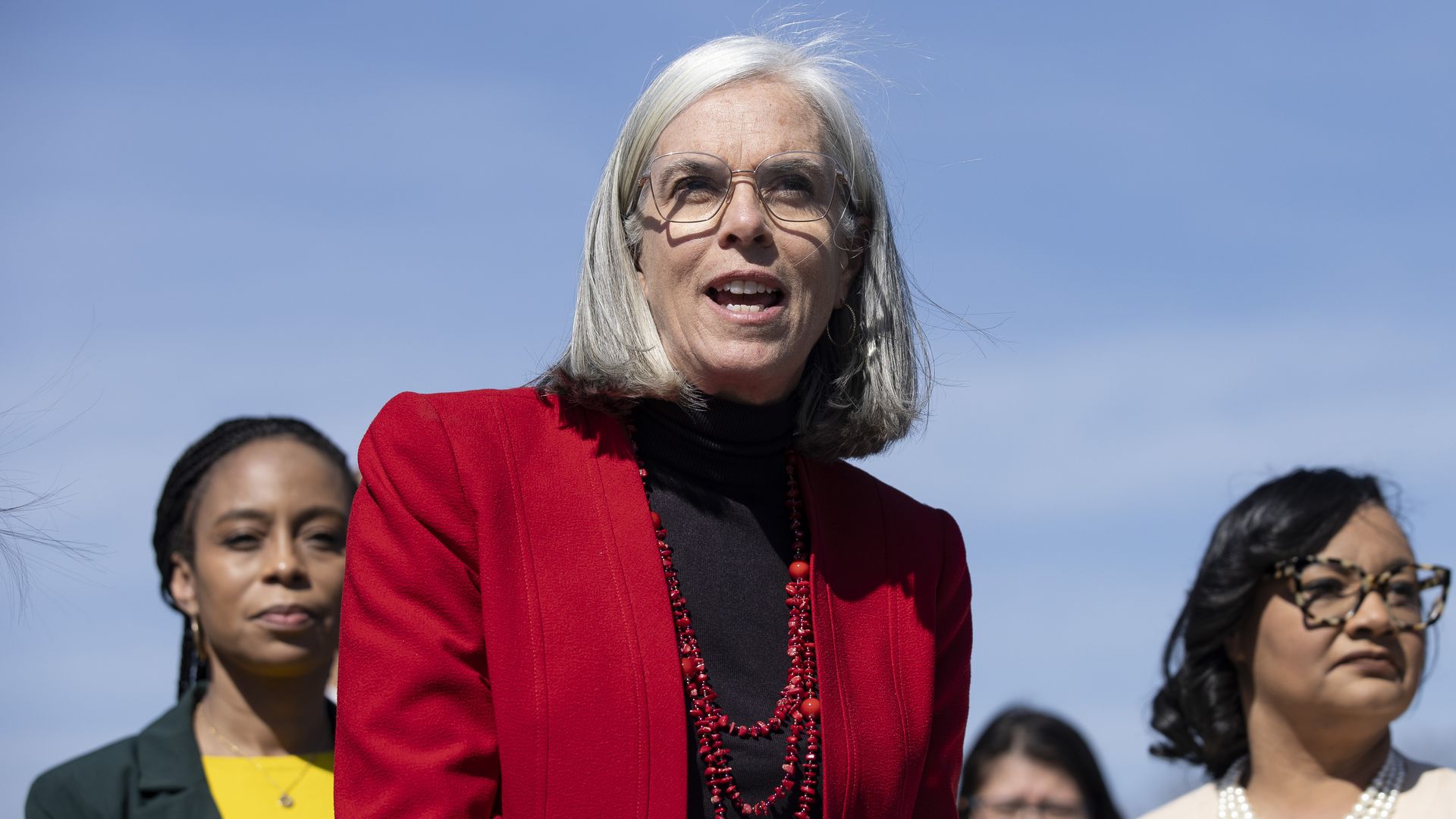 Rep. Katherine Clark, wearing a red blazer and black shirt, surrounded by colleagues under a blue sky.
