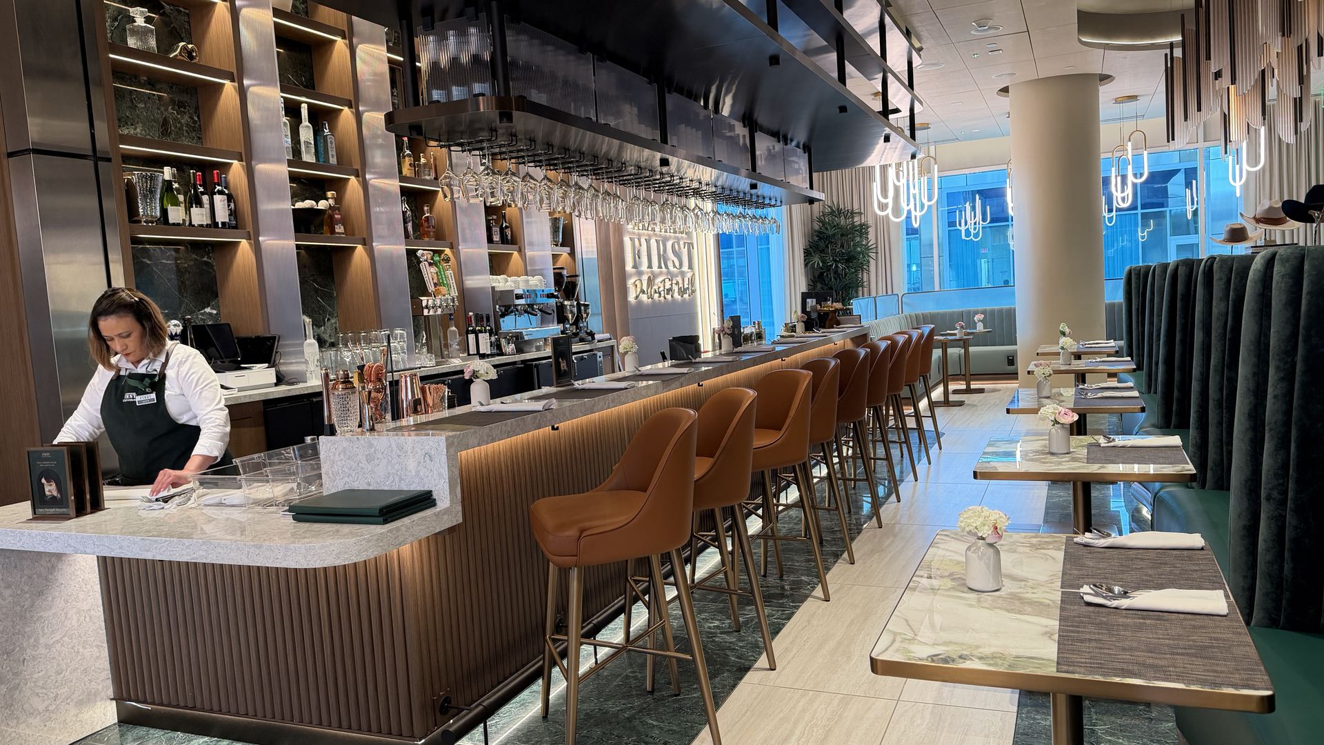Modern bar interior with a marble counter, copper stools, bottle shelves, hanging wine glasses, a bartender in white and green, warm lighting, and blue seating by large windows.