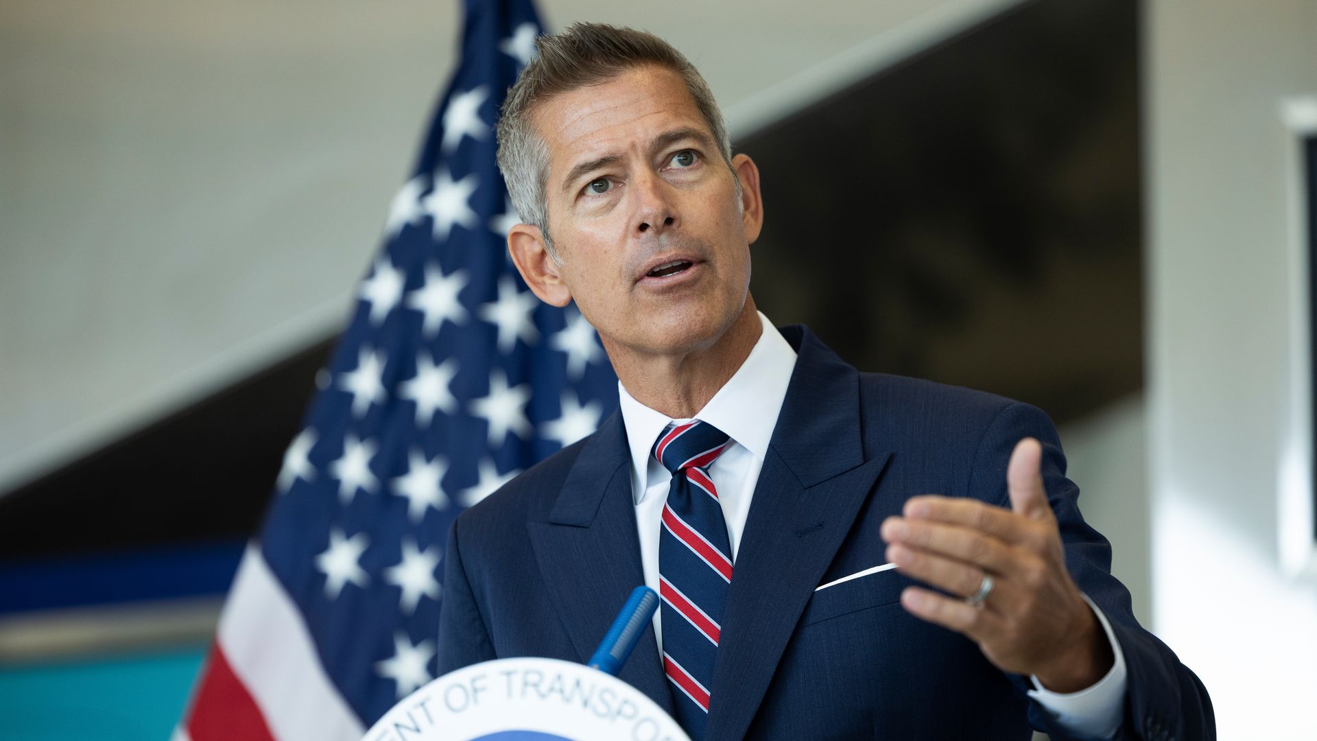 Sean Duffy — wearing a navy blue suit, a white collared shirt and a striped tie — gestures as he speaks in front of an American flag.