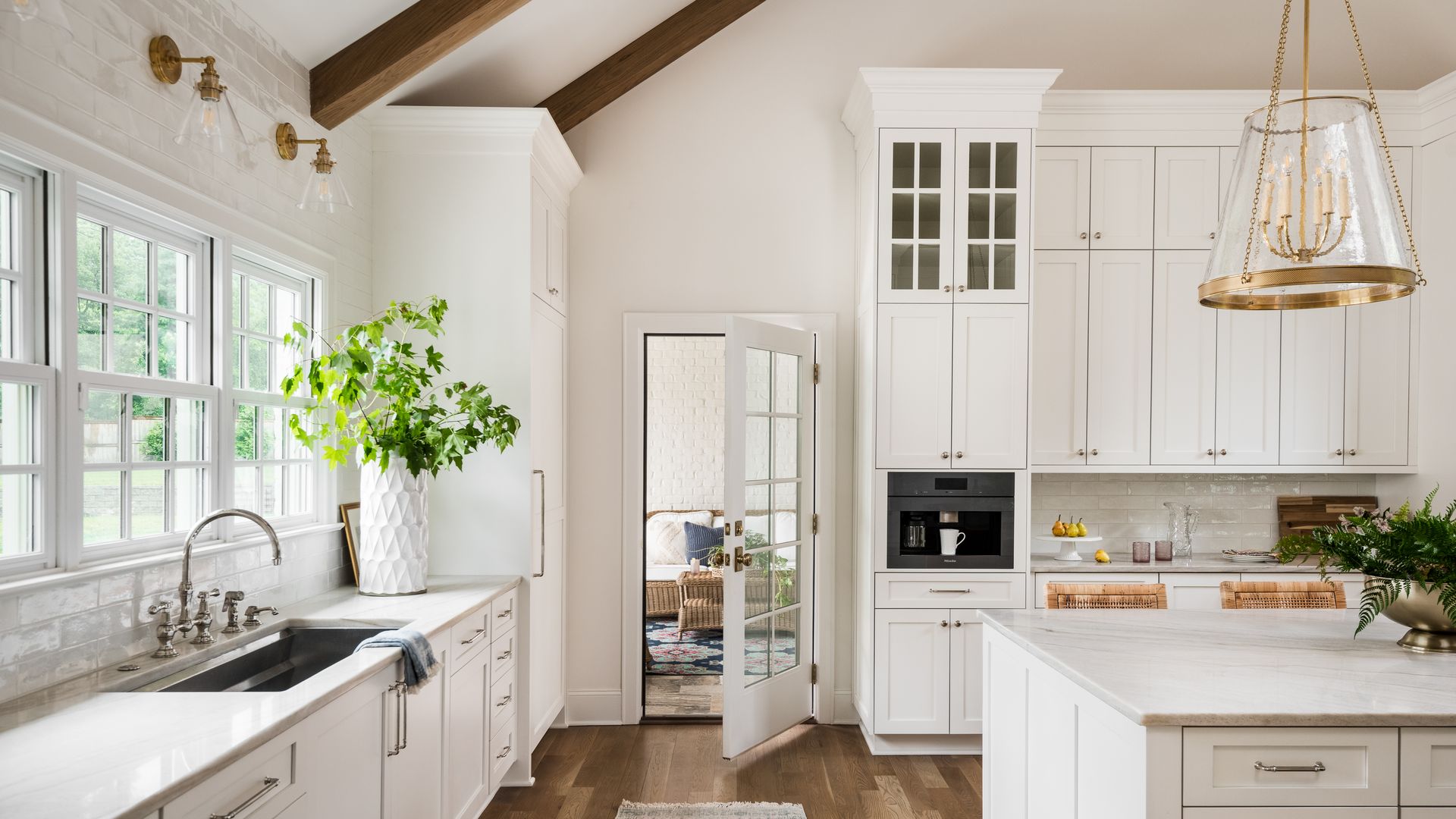 Bright white kitchen with wooden floors, large windows, green plant in white vase, glass door leading to living room, marble countertops, and brass light fixture above island.