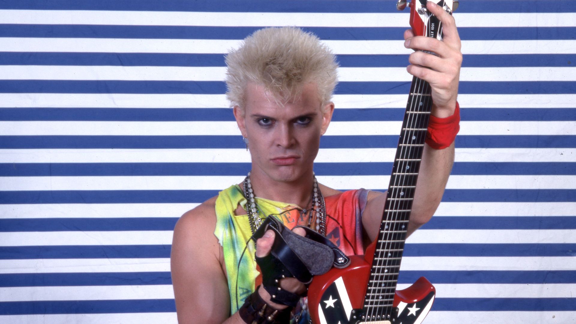 Punk rocker with bleached spiky hair, wearing a colorful tie-dye top and necklaces, grips a red electric guitar with star decals against a blue-and-white striped wall.