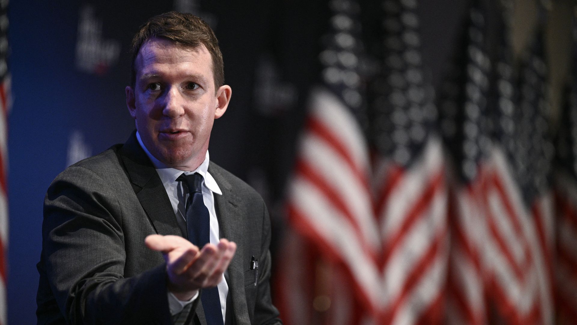 A person in a suit gestures with their hand in front of American flags