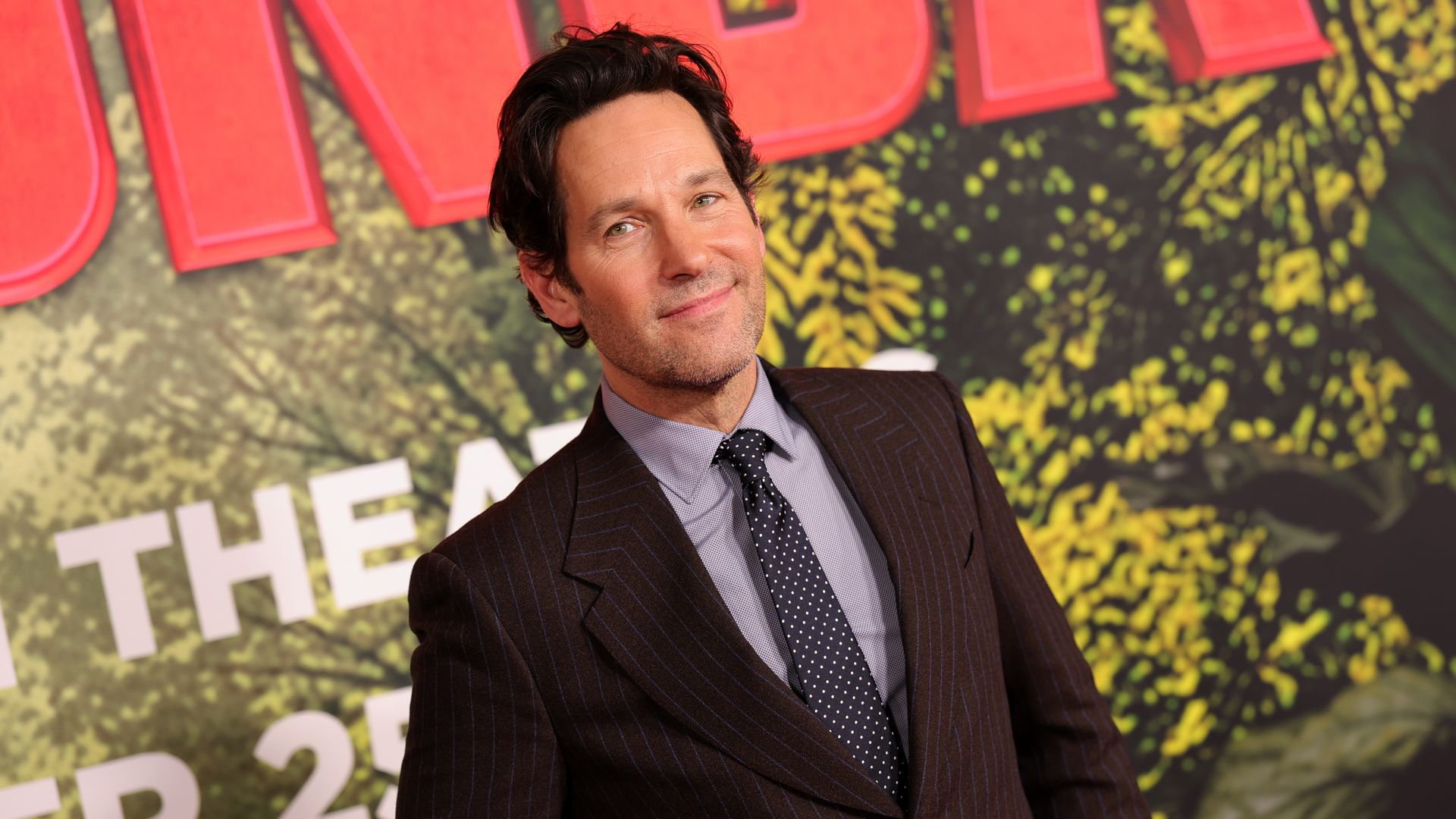 Man with dark wavy hair in a dark pinstripe suit and polka dot tie stands smiling in front of a colorful movie premiere backdrop with red and yellow text and foliage.