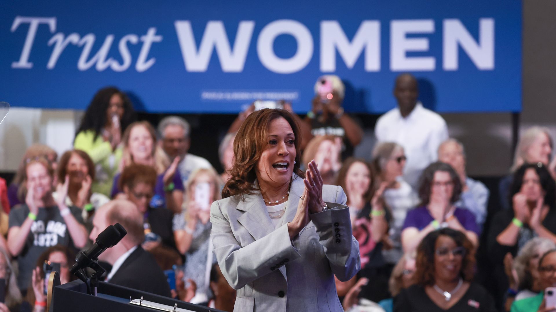 A woman in a gray suit clapping her hands with a group of about a dozen people behind her on risers below a blue sign that reads "Trust WOMEN" in white letters.