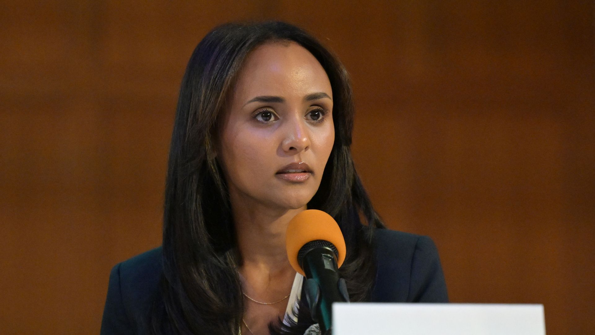 Close-up of a woman with long dark hair in a navy blazer speaking into an orange microphone at a panel; a nameplate in front reads 'Melat Kiros' against a brown backdrop.