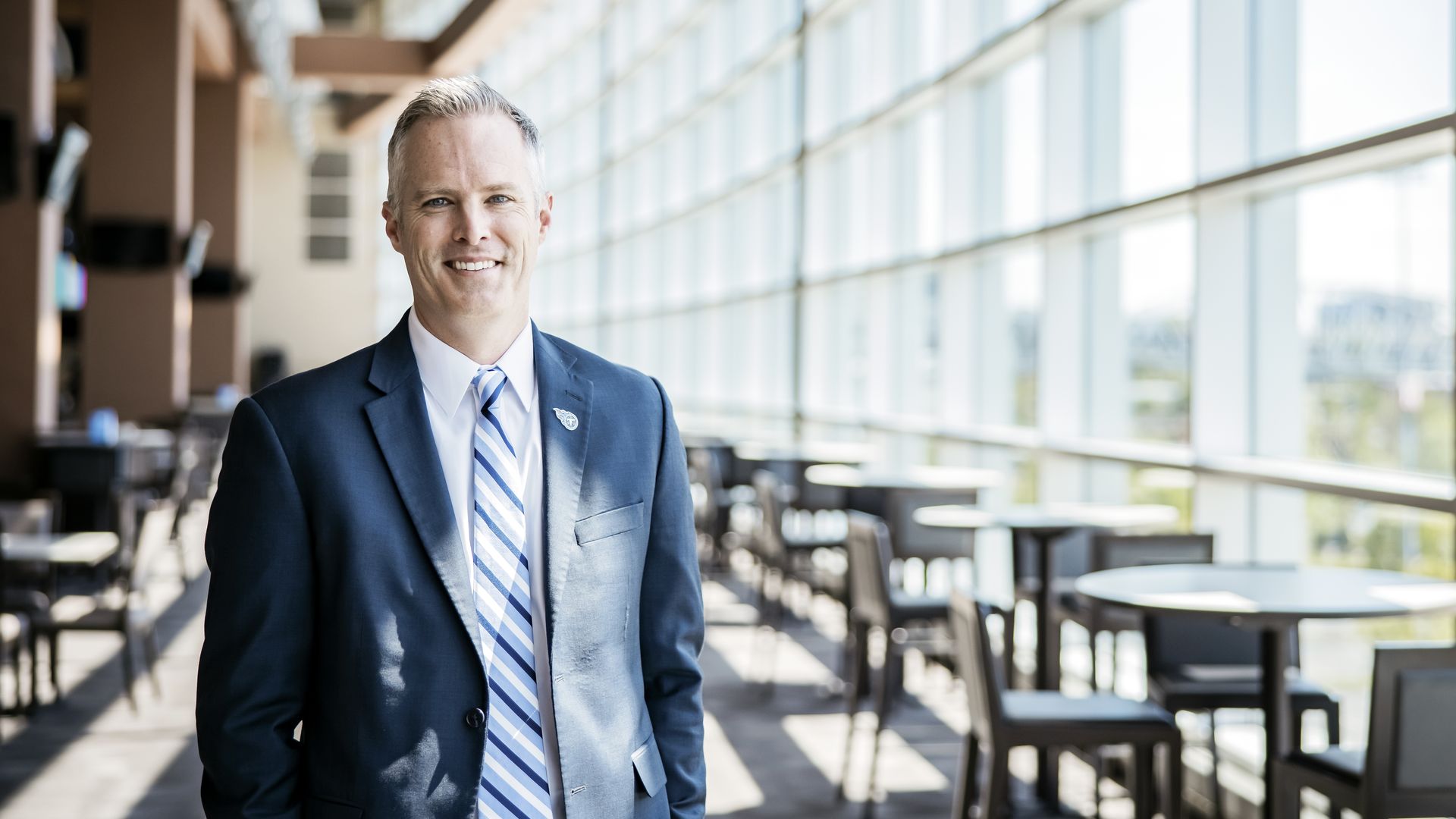 Smiling man in a navy suit and striped tie stands in a bright, modern atrium with large glass walls and tables and chairs in the background.