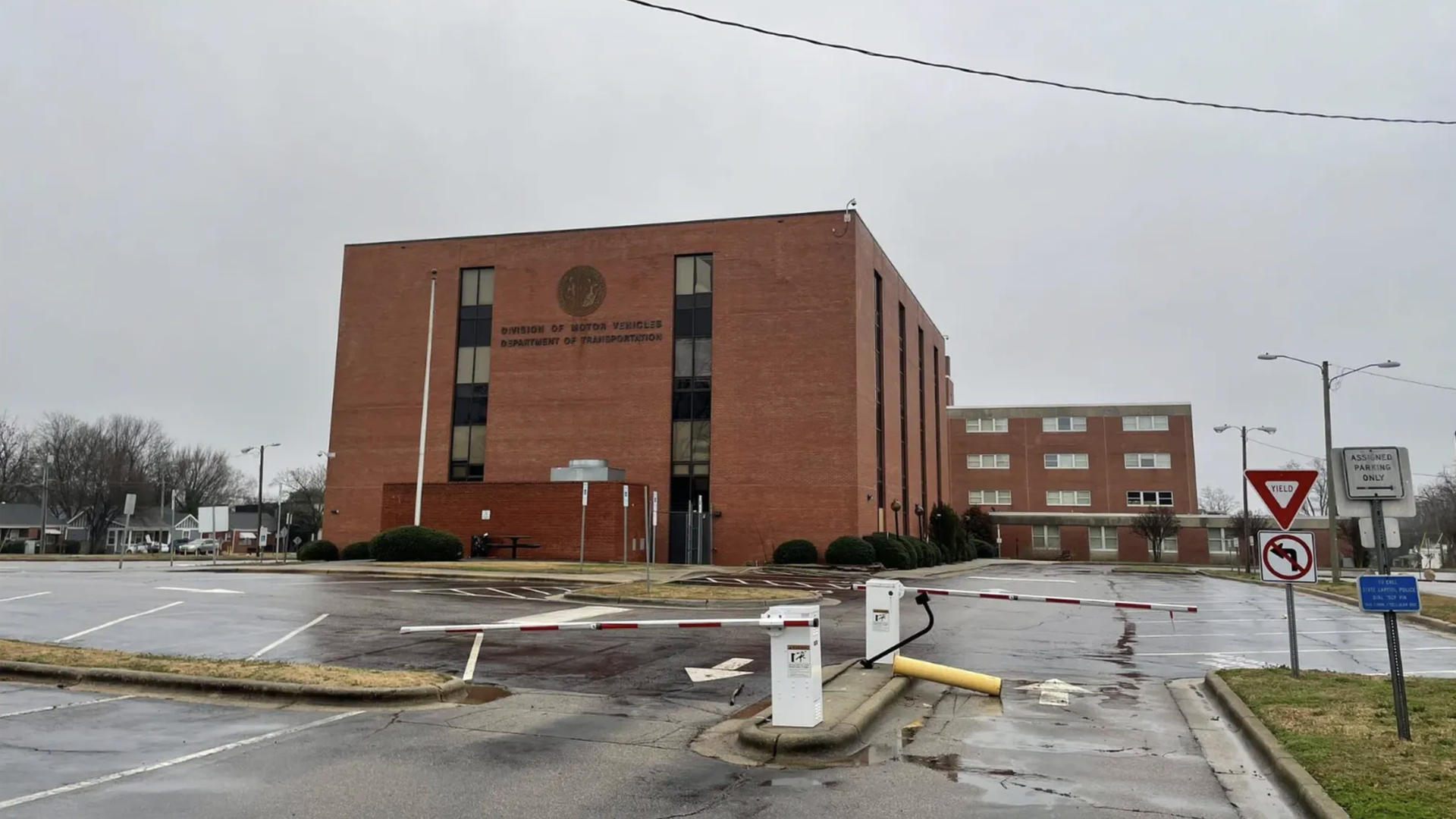 Red brick government building with a circular emblem and the Division of Motor Vehicles sign on the facade. A parking lot with barriers and road signs, including a yield sign on the right.