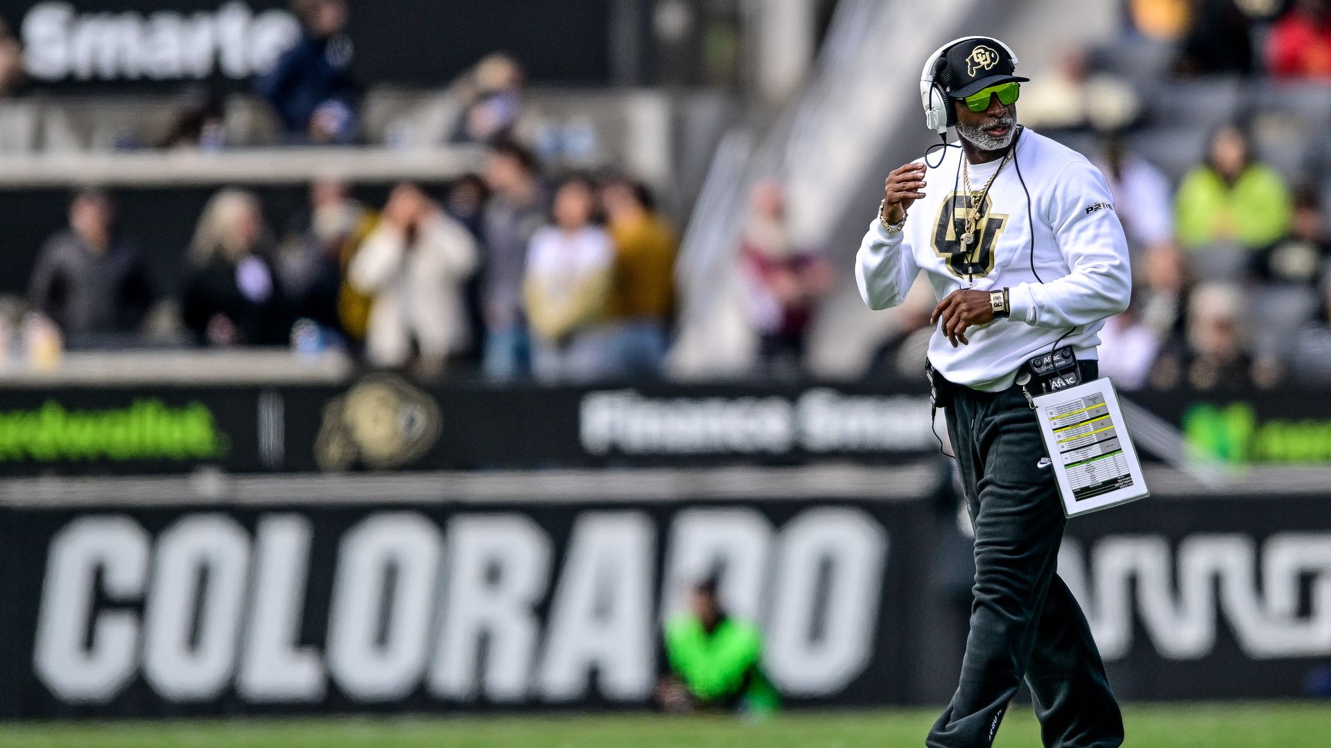 An older football coach on a green field, wearing a white Colorado sweatshirt, black pants, headset, sunglasses, and a clipped play sheet; blurred stands and a COLORADO sign behind him.