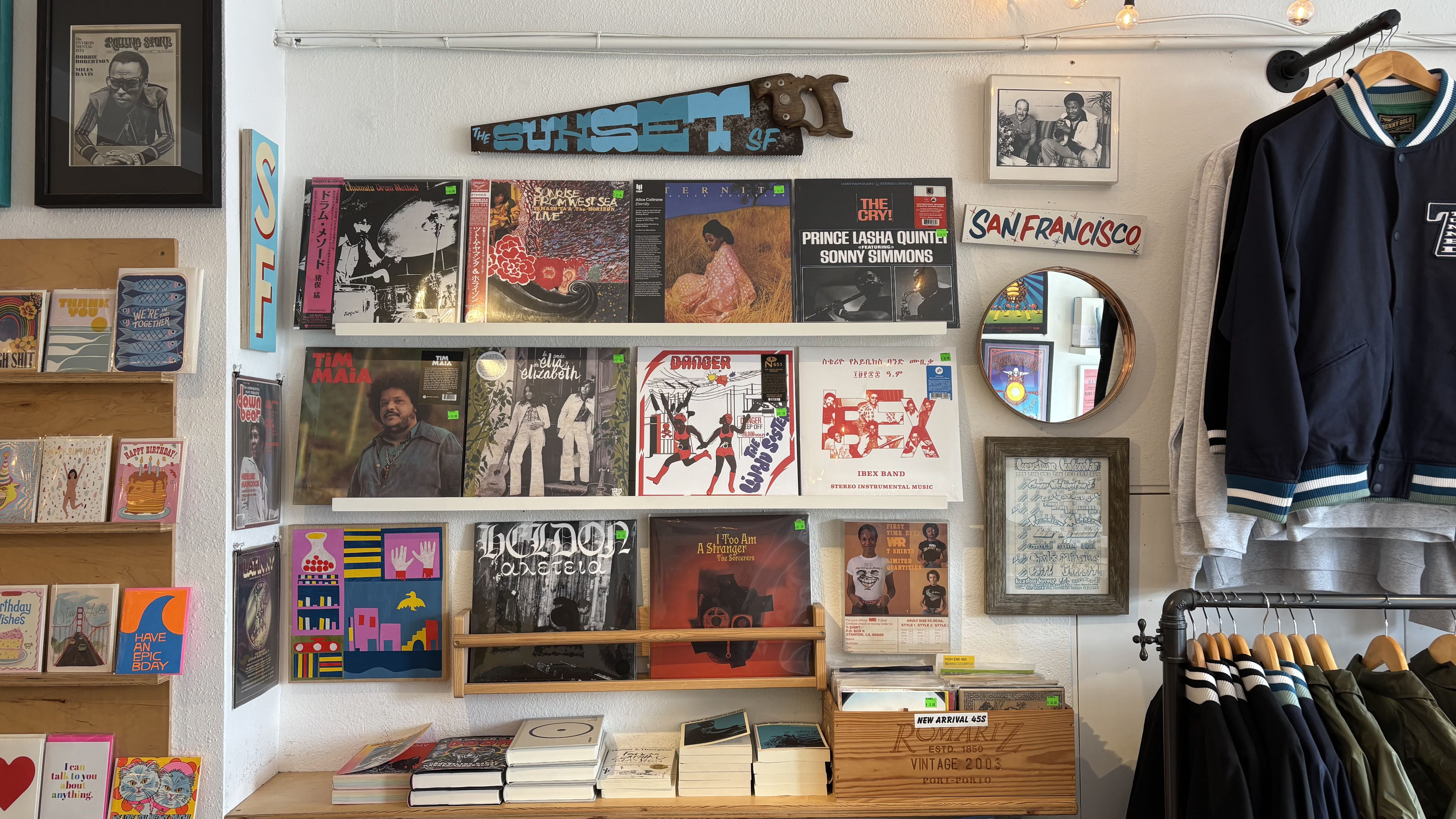 Interior of a record store wall with jazz and soul vinyl records displayed on shelves, a round mirror, San Francisco sign, framed photos, greeting cards on left, and jackets on hangers on right.