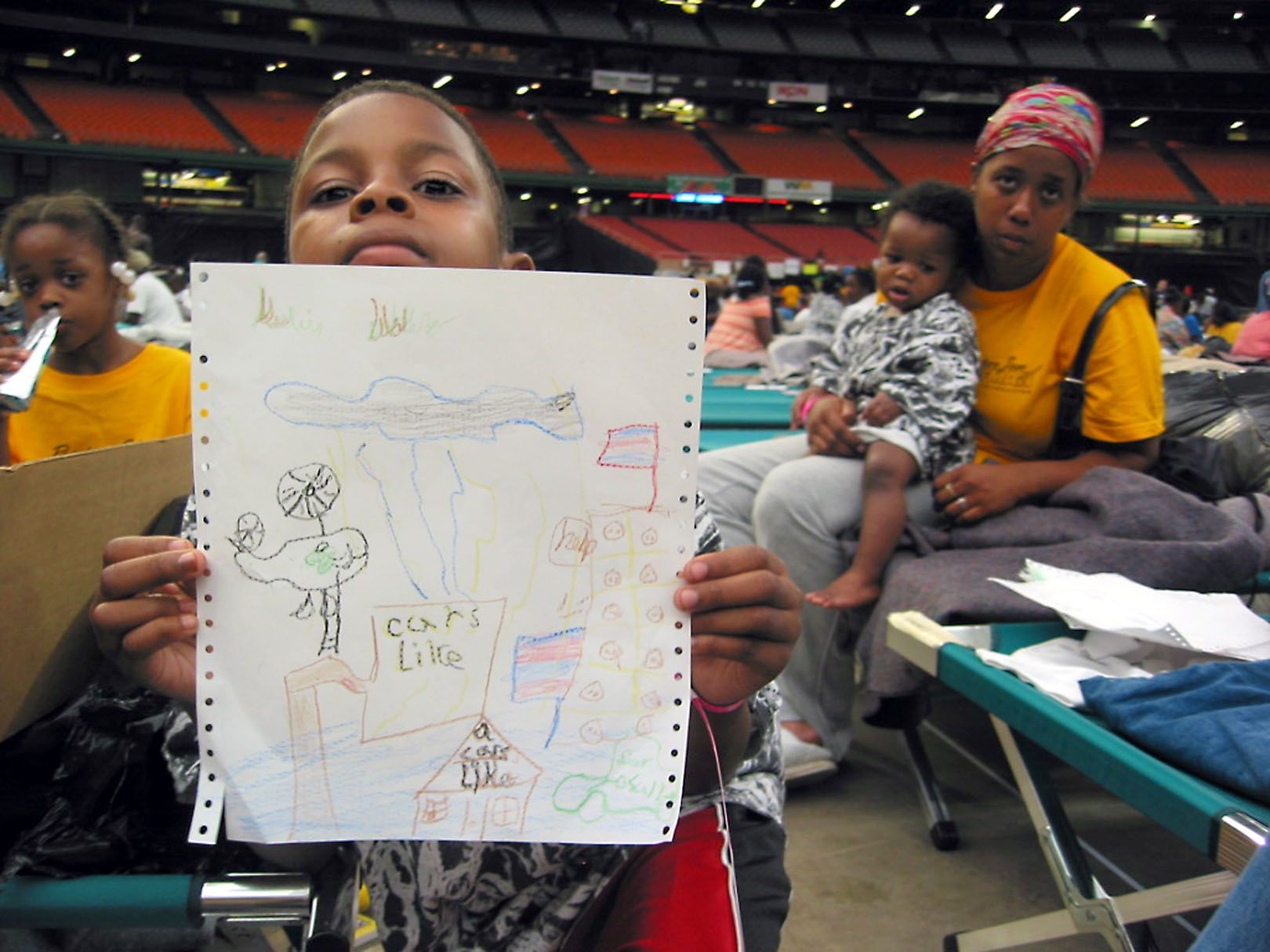 A boy in a shelter holds up a colorful drawing featuring storms, helicopters, buildings, and flags.