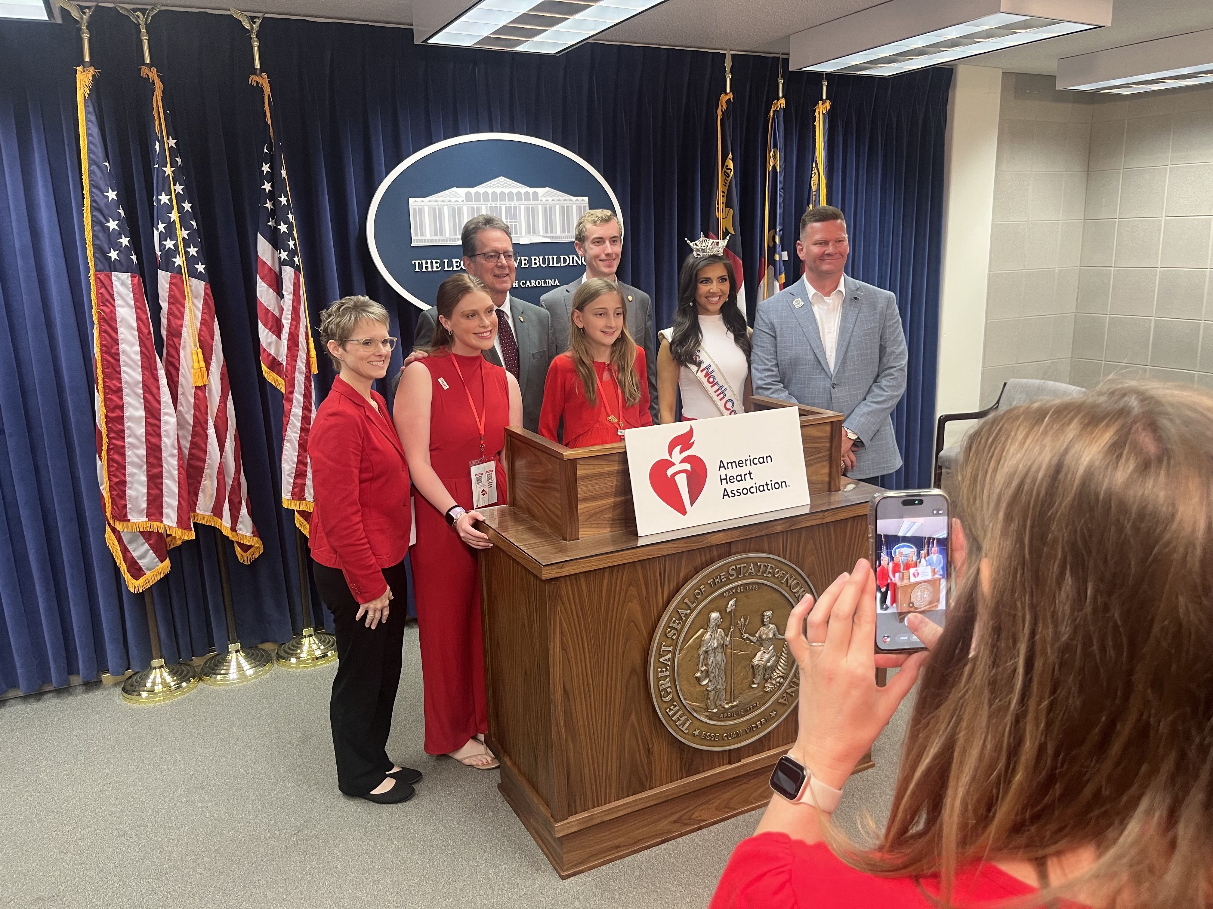 Group photo at a North Carolina state Capitol podium: blue curtains, several American flags, a crowned woman in a sash among others in red. A bystander take a photo with a phone; podium shows American Heart Association.