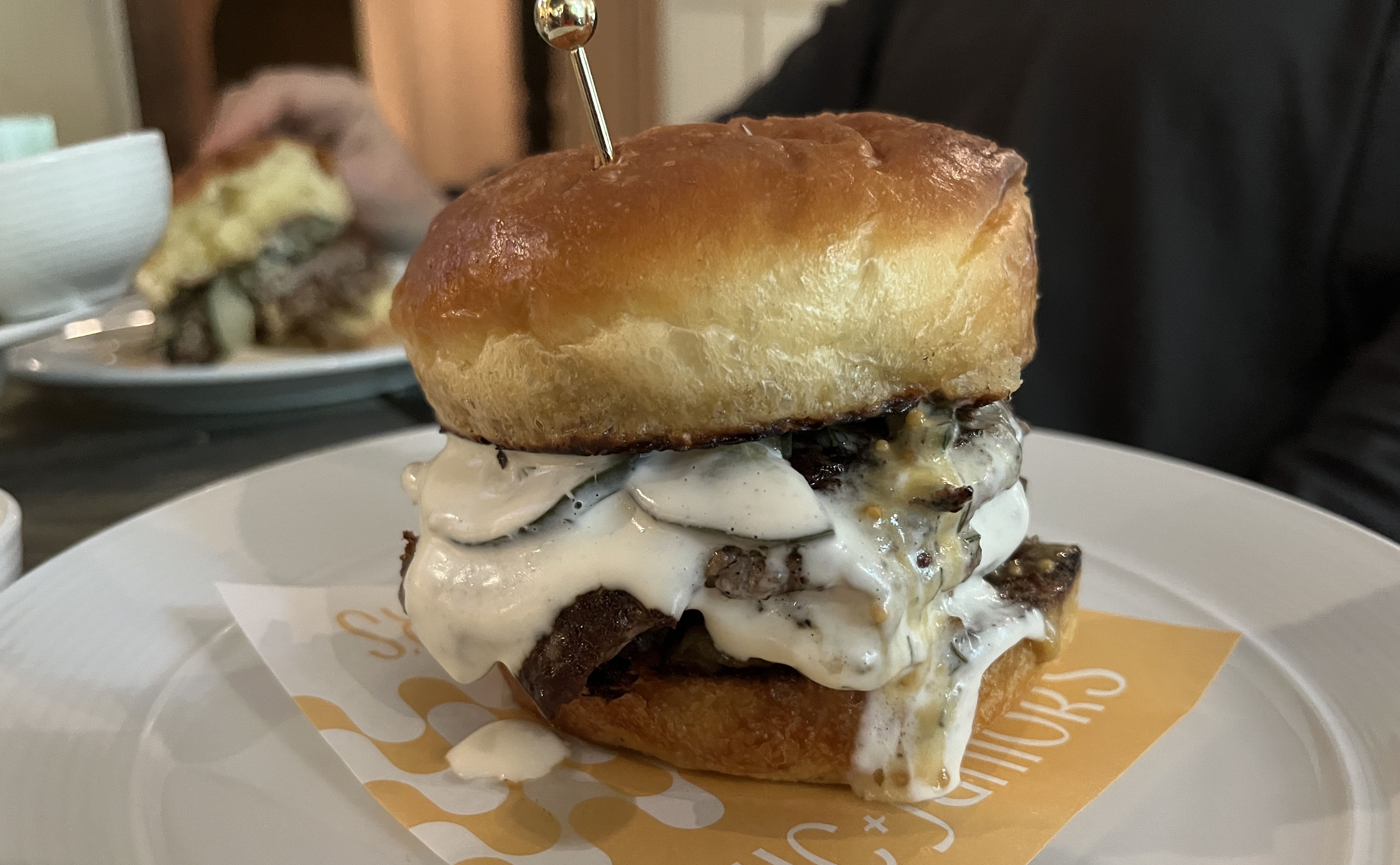 Close-up of a juicy cheeseburger with melted white cheese sauce dripping over a toasted golden-brown bun, served on a white plate with orange paper. A person holds another burger in the background.