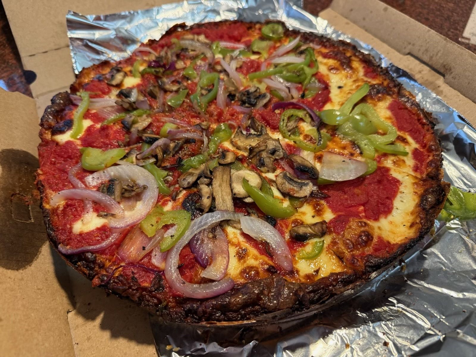 Charred-crust pizza in a cardboard box with foil lining. Topped with red sauce, melted cheese, green bell peppers, onions, mushrooms, and other vegetables.